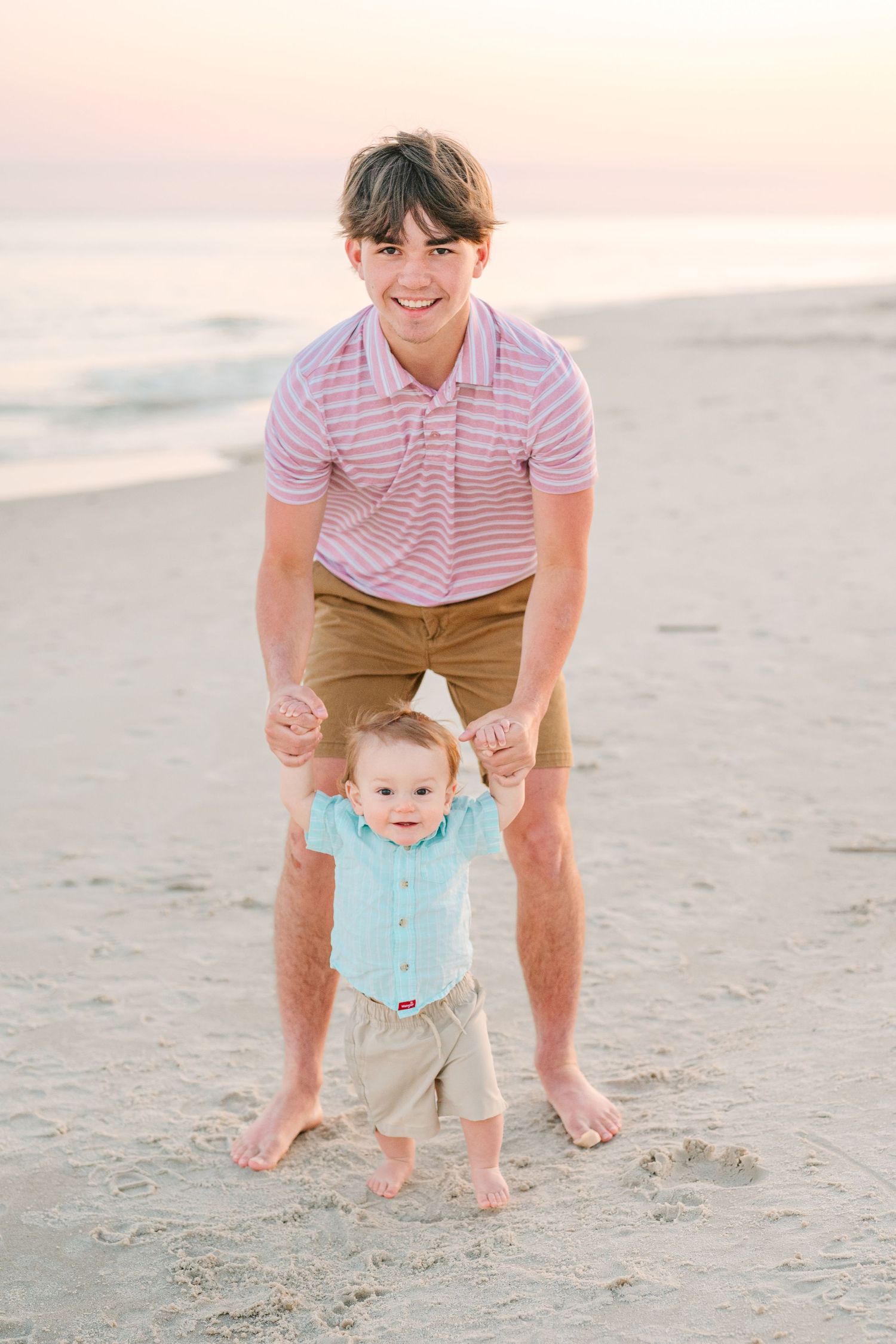 A touching moment captured on the beach at sunset with someone helping a baby take their first steps in the sand.