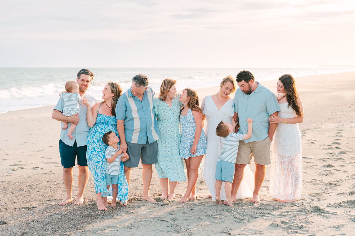 Large family group wearing coordinated blue and white outfits stands together on a sandy beach.