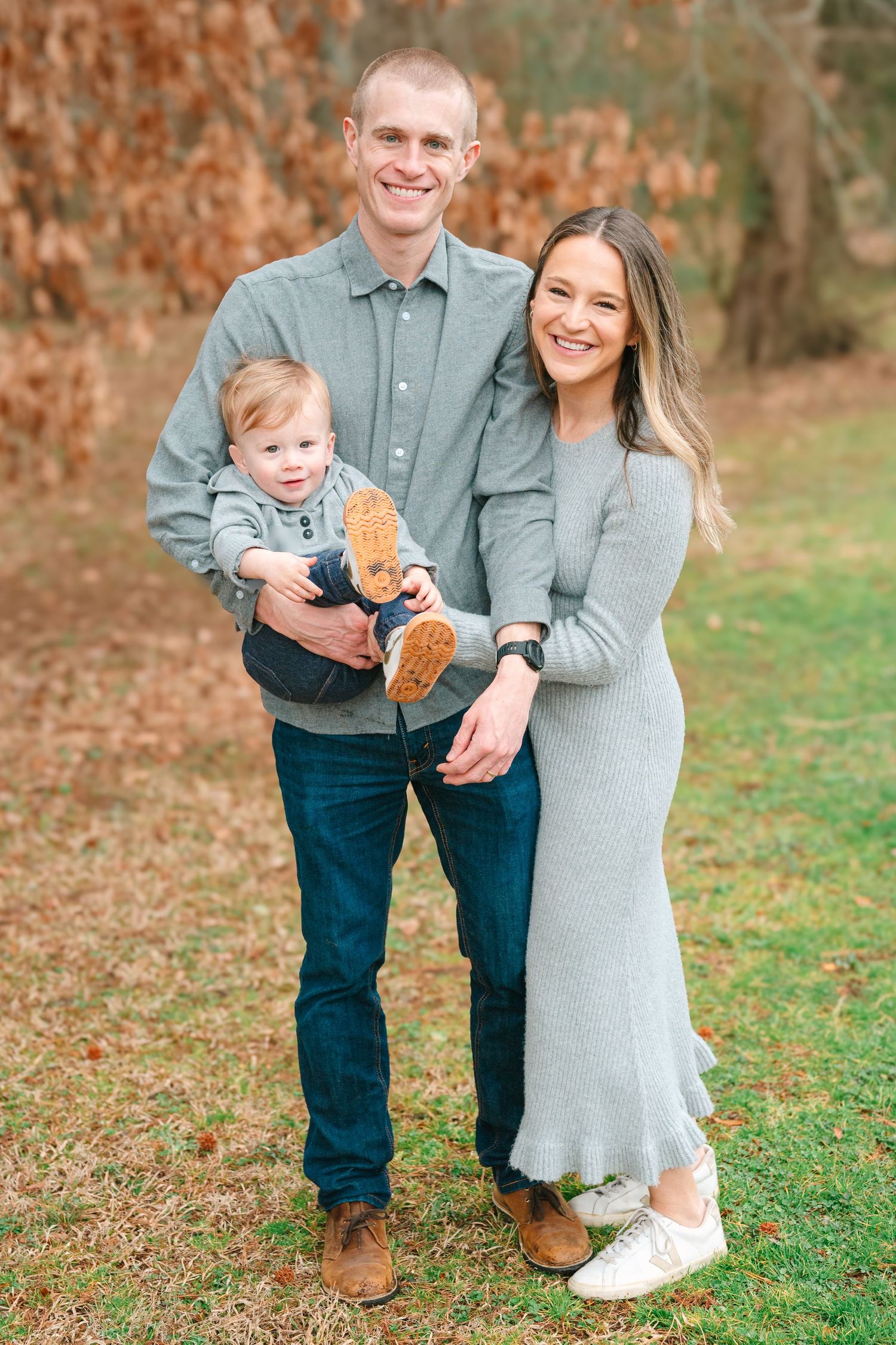 A young family poses outdoors in coordinating gray outfits against fall foliage, with their small child holding a stuffed toy.