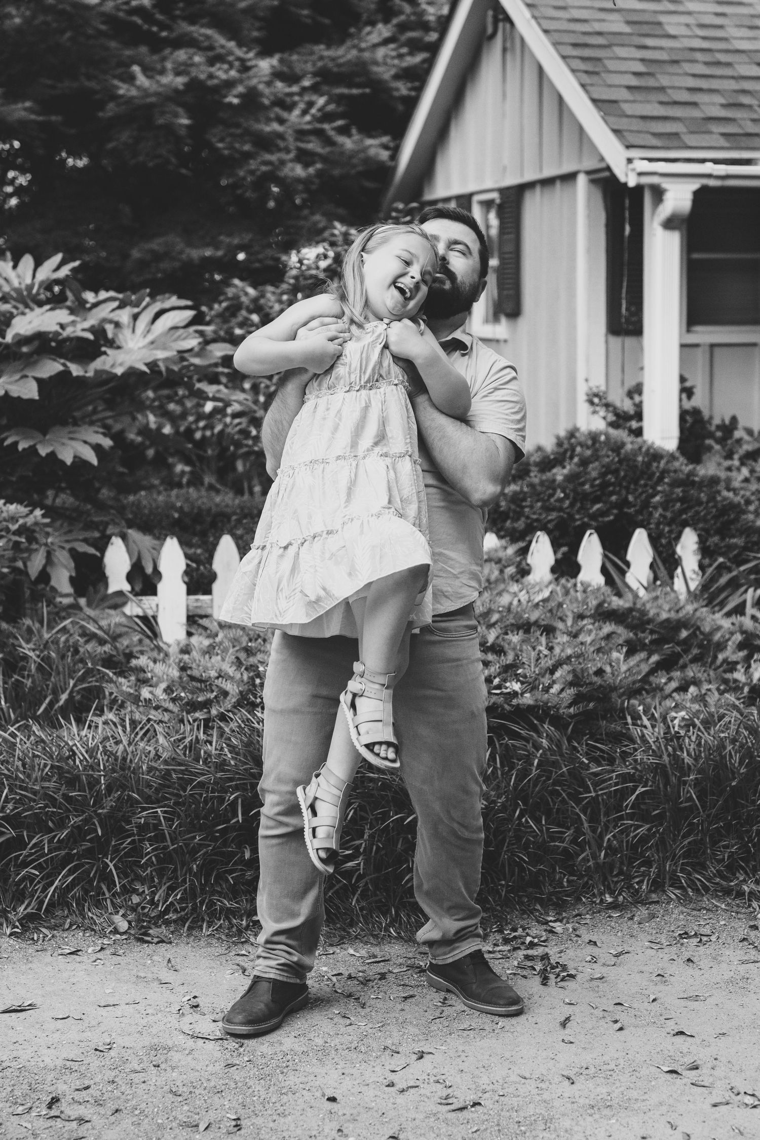 A joyful black and white photo of a playful moment shared between parent and child in front of a cottage with white fence.
