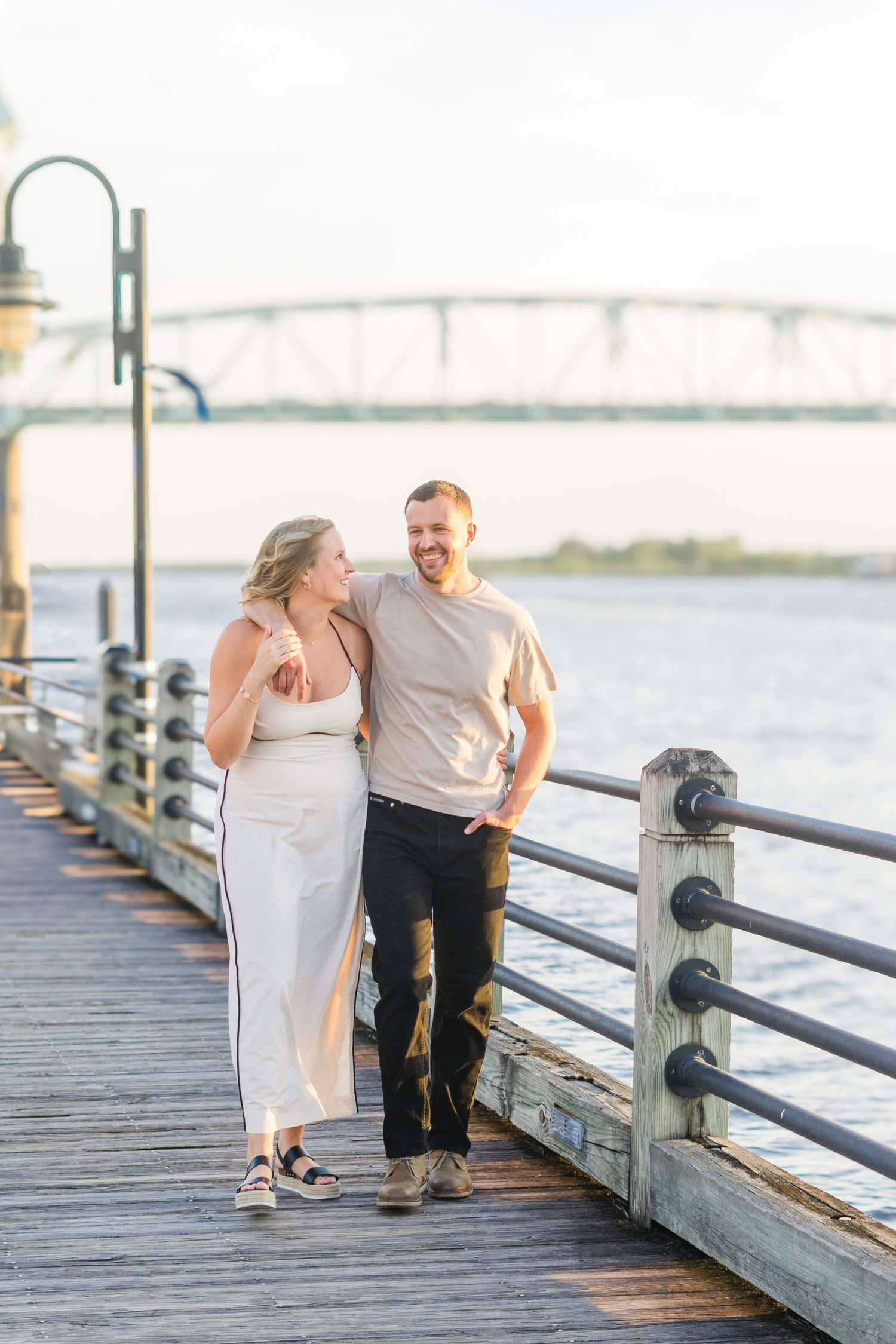 A couple takes a romantic stroll along a wooden pier with a bridge visible in the background at sunset.
