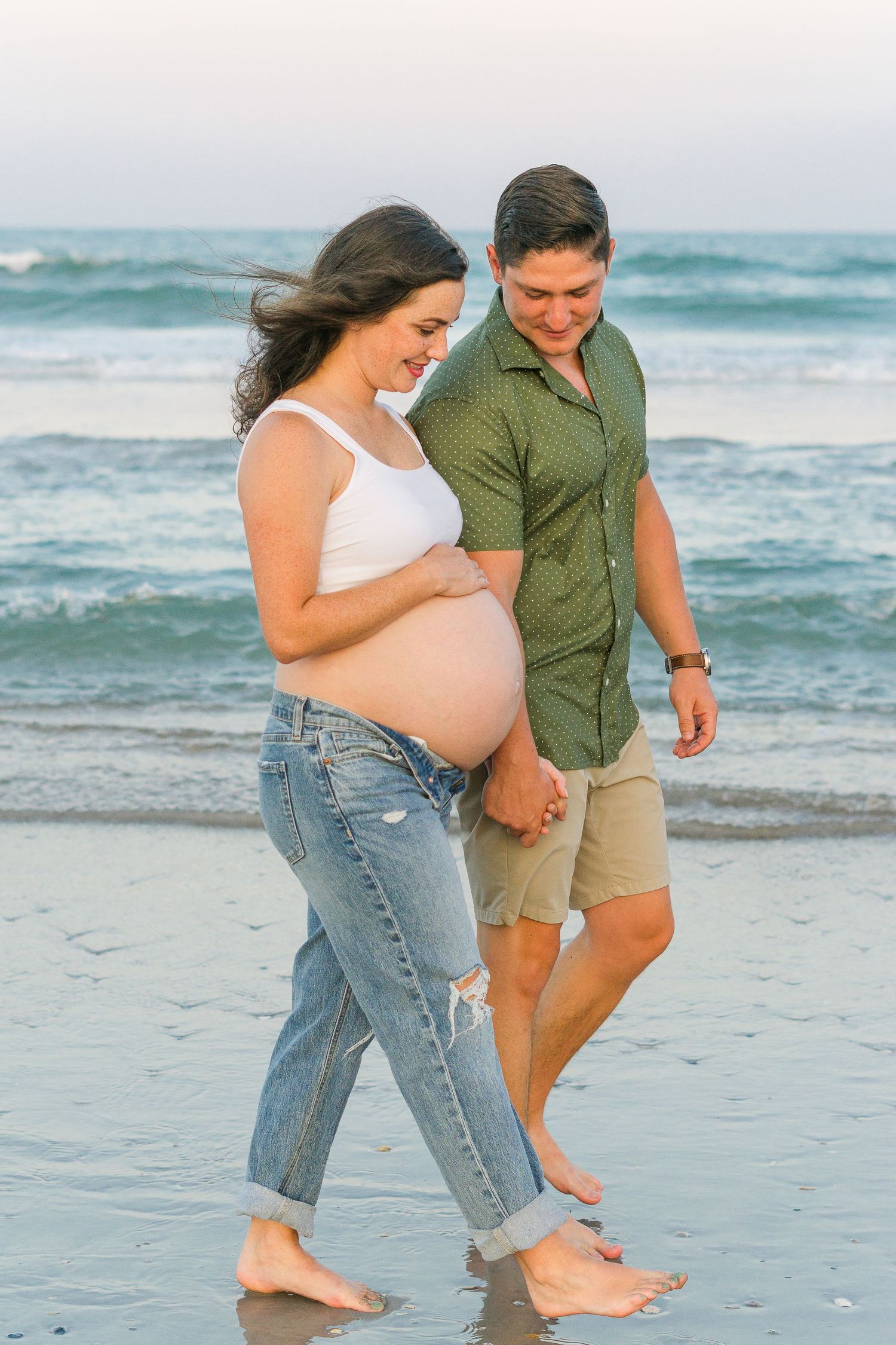 Casual beach maternity shoot with couple walking along shoreline.
