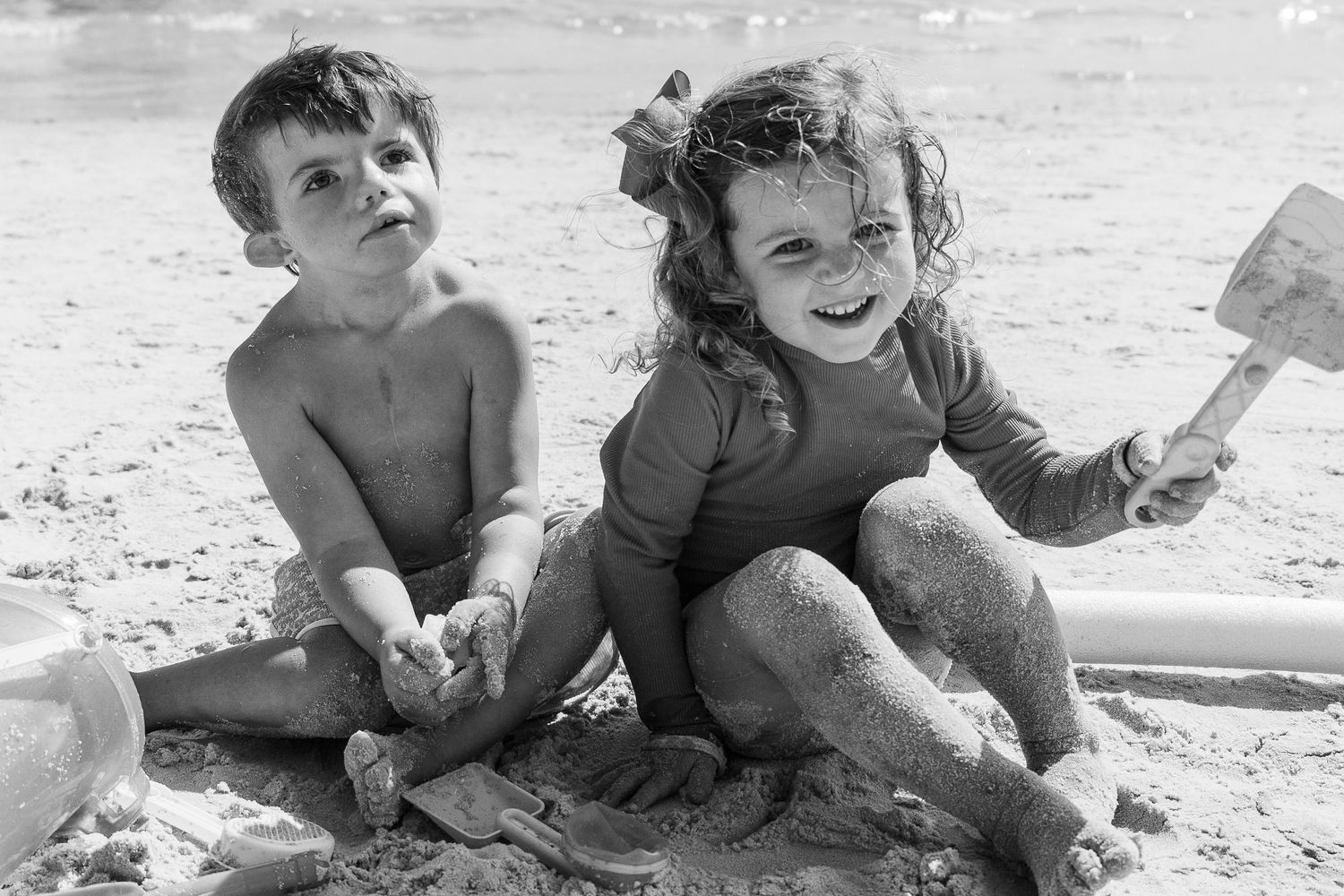 Two children playing joyfully in the sand at the beach in a nostalgic black and white photograph.