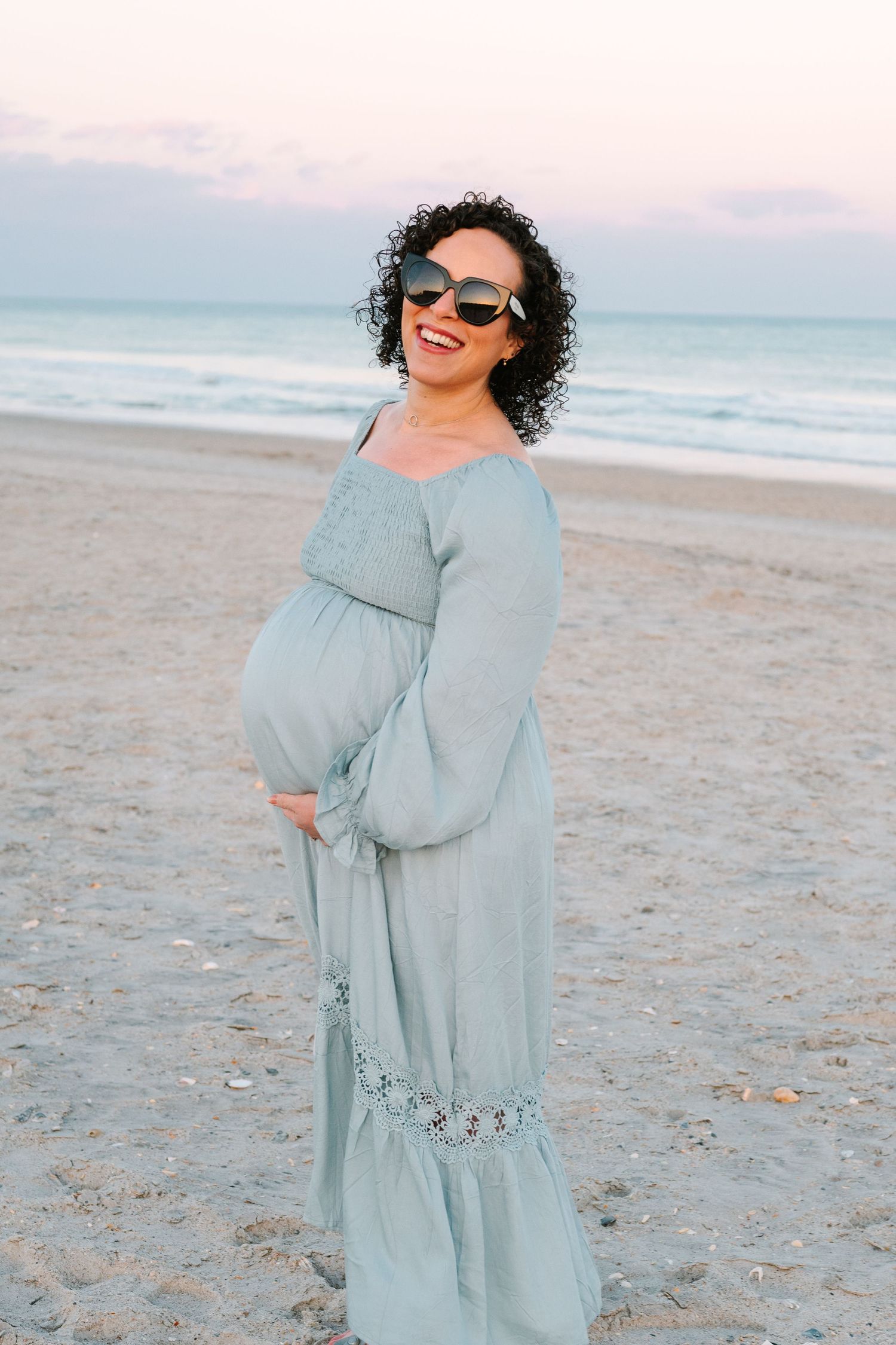A pregnant person in a flowing blue dress and sunglasses smiles while standing on a beach at sunset.