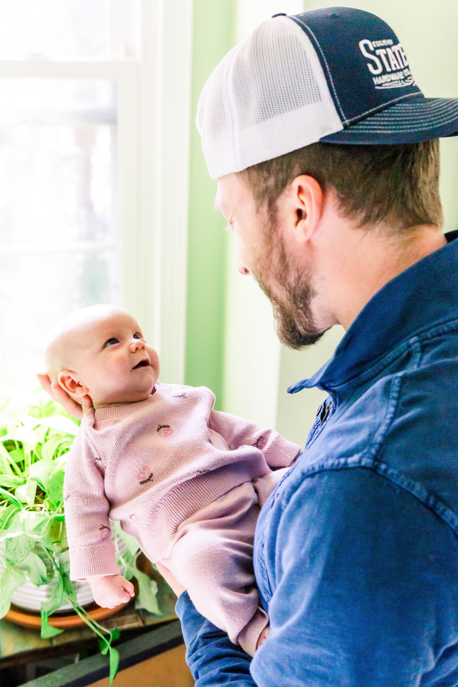 A baby in pink clothing smiles up adoringly at their dad wearing a baseball cap and blue jacket.