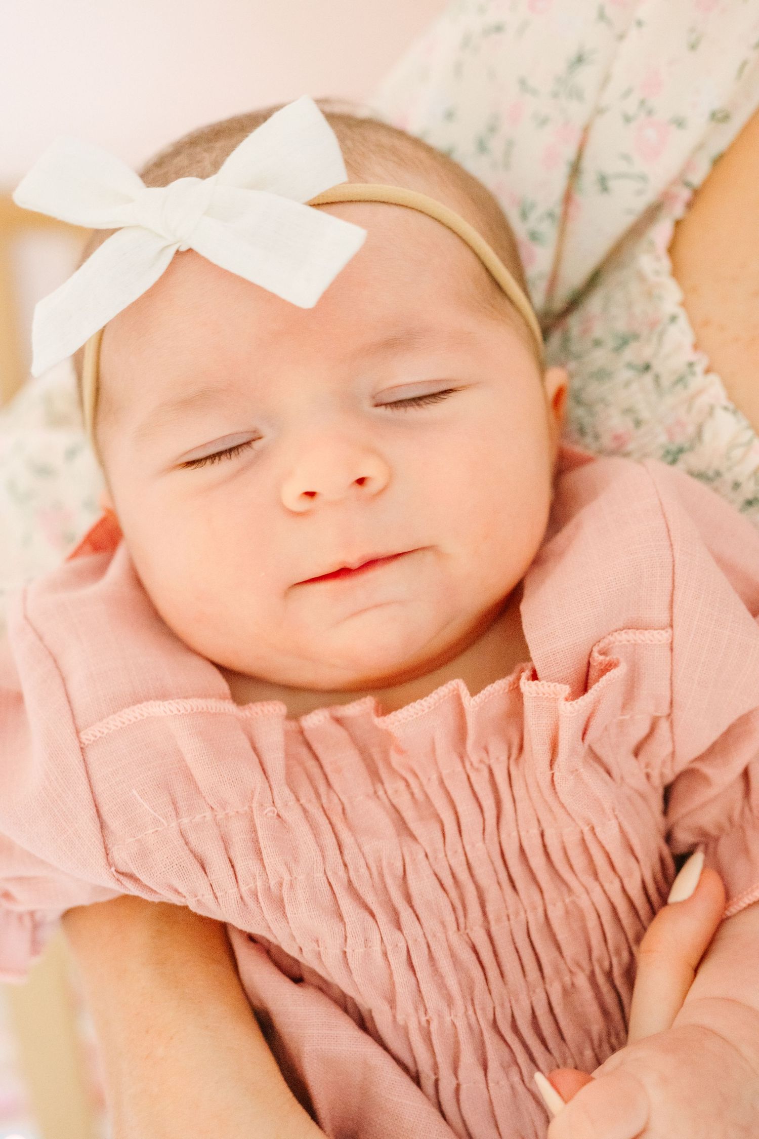 A sleeping baby wearing a pink smocked dress and white bow headband rests peacefully.