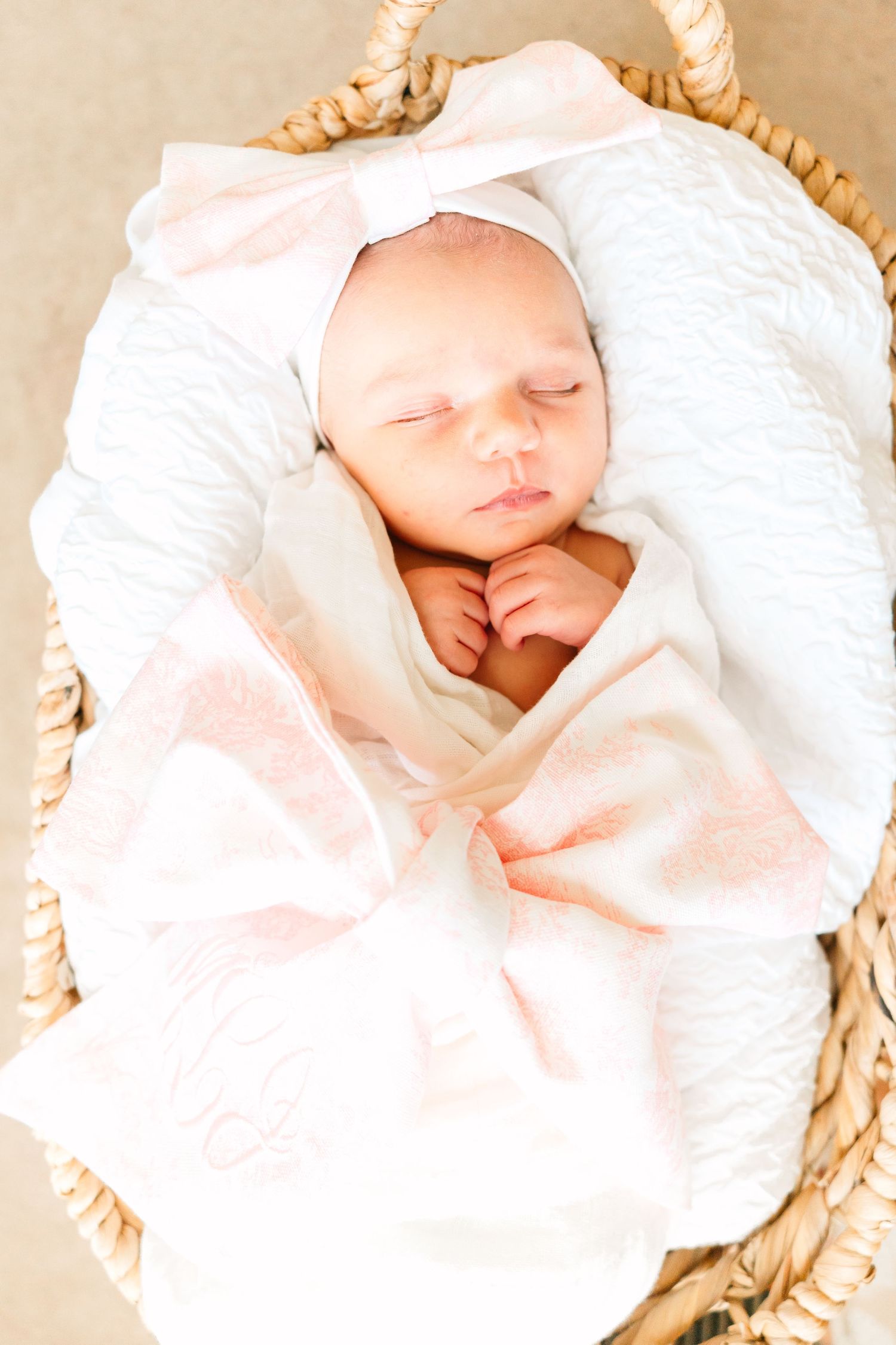 A newborn sleeping peacefully in a woven basket wrapped in soft white and pink blankets with a bow.