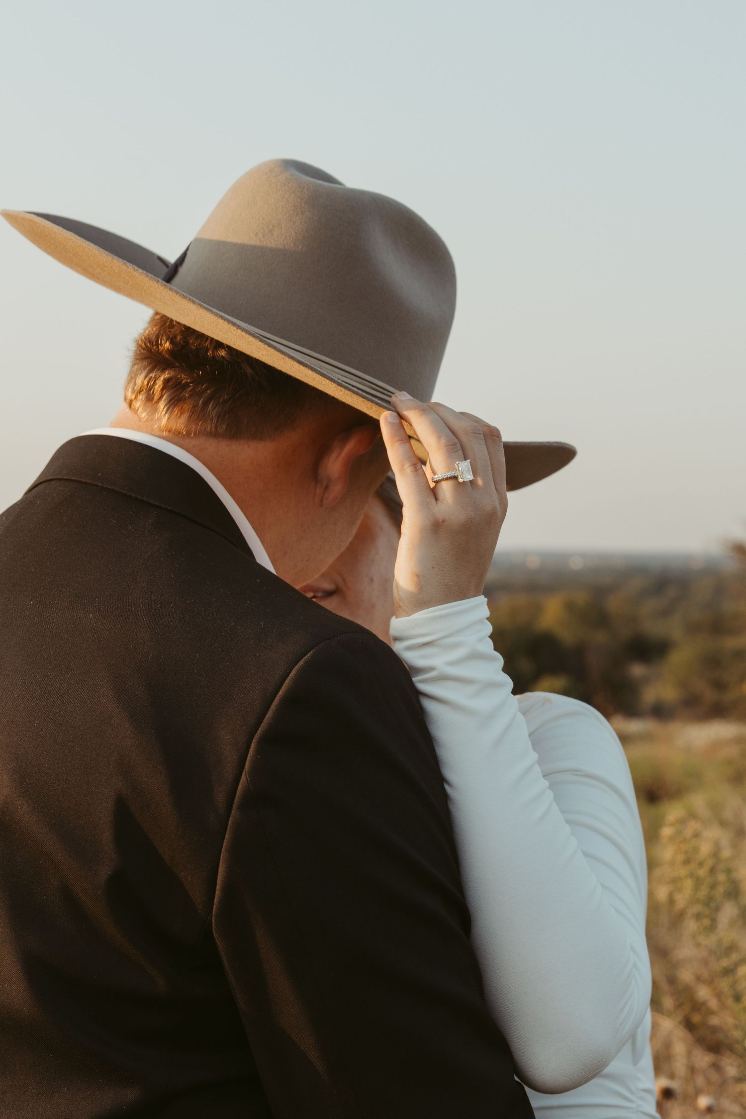 A person in western attire wearing a brown cowboy hat adjusts their brim against a sunset backdrop.
