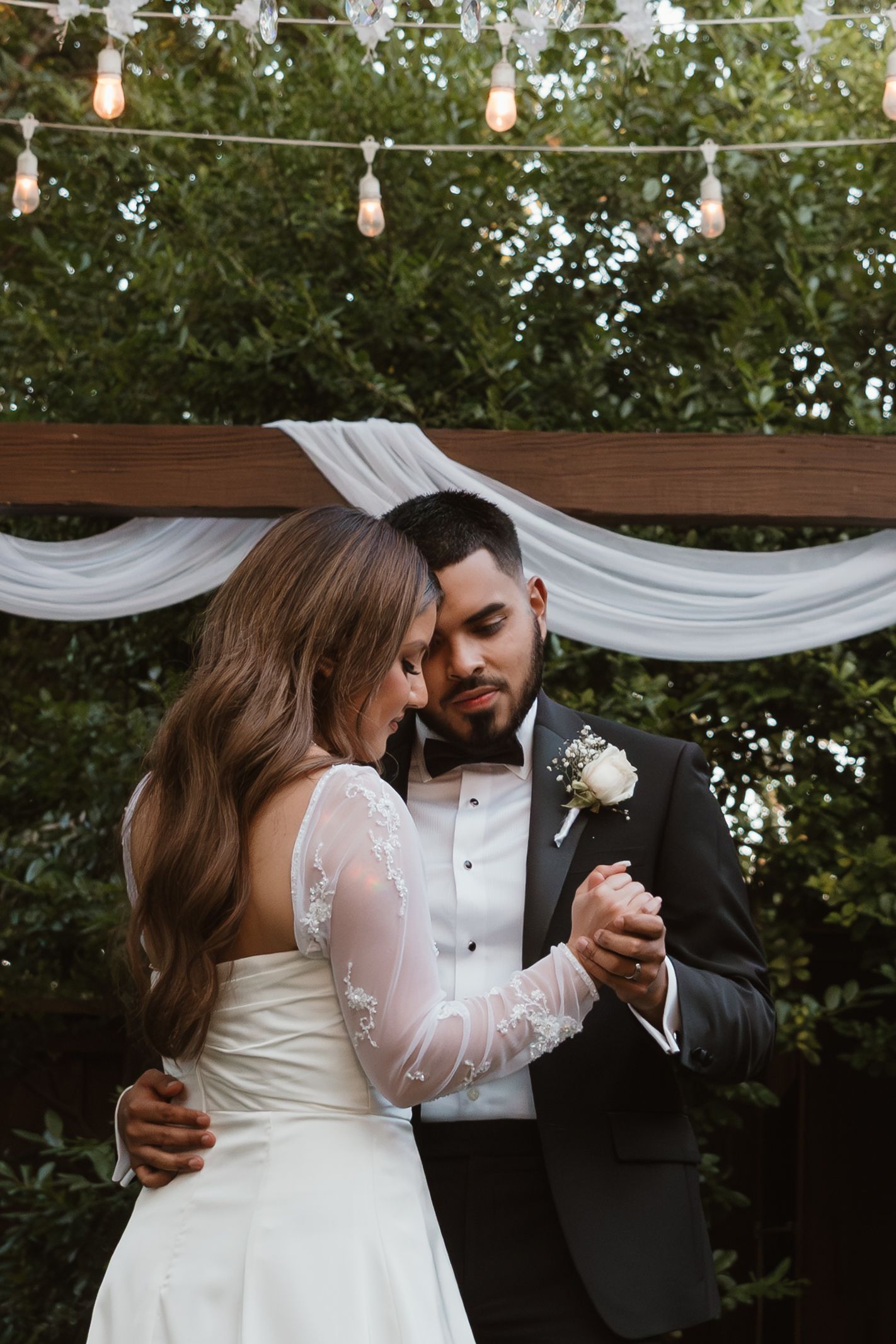 Romantic outdoor wedding scene with couple embracing under string lights and white fabric draping.