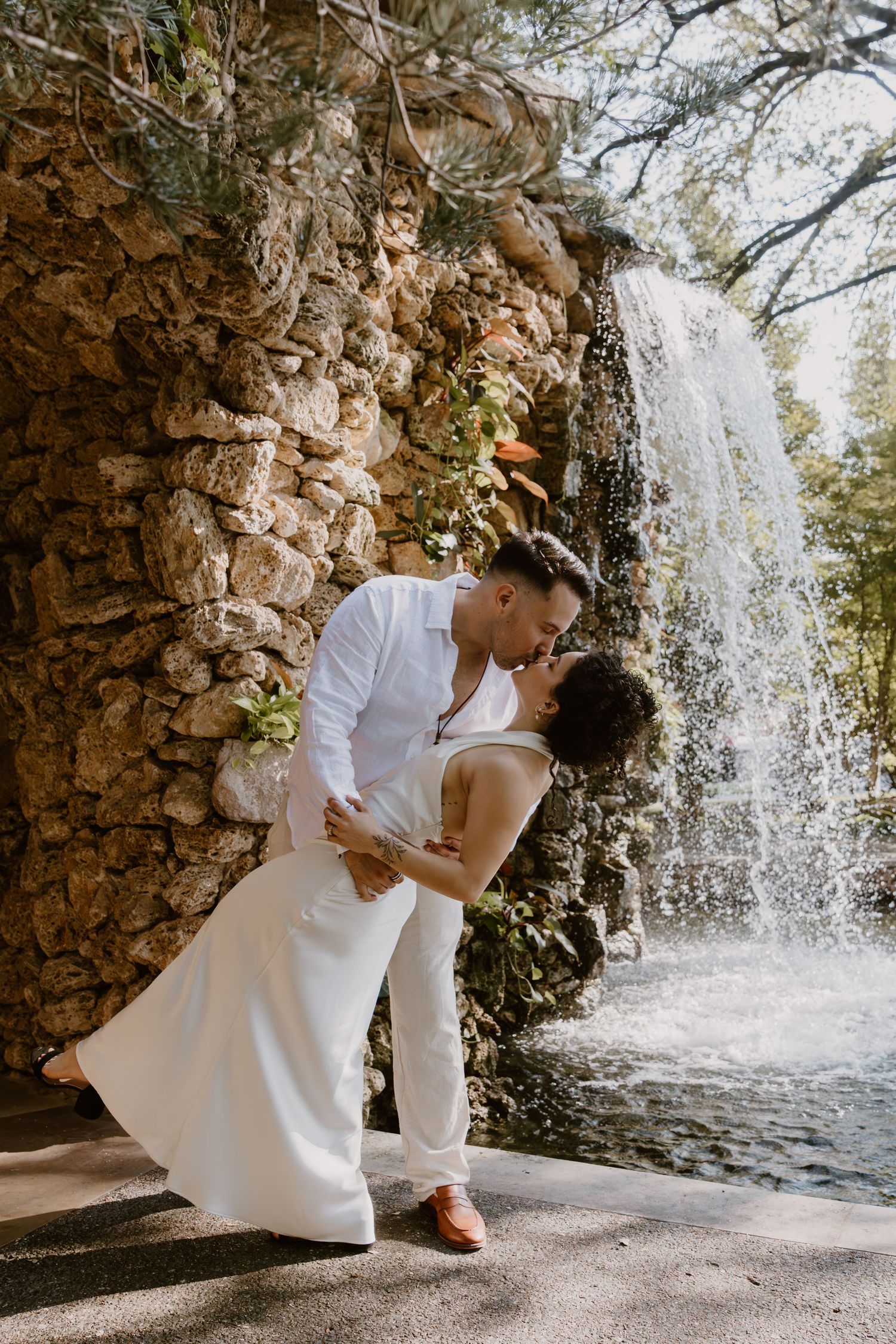 A romantic couple shares intimate moments in front of a cascading waterfall in a scenic stone grotto setting.