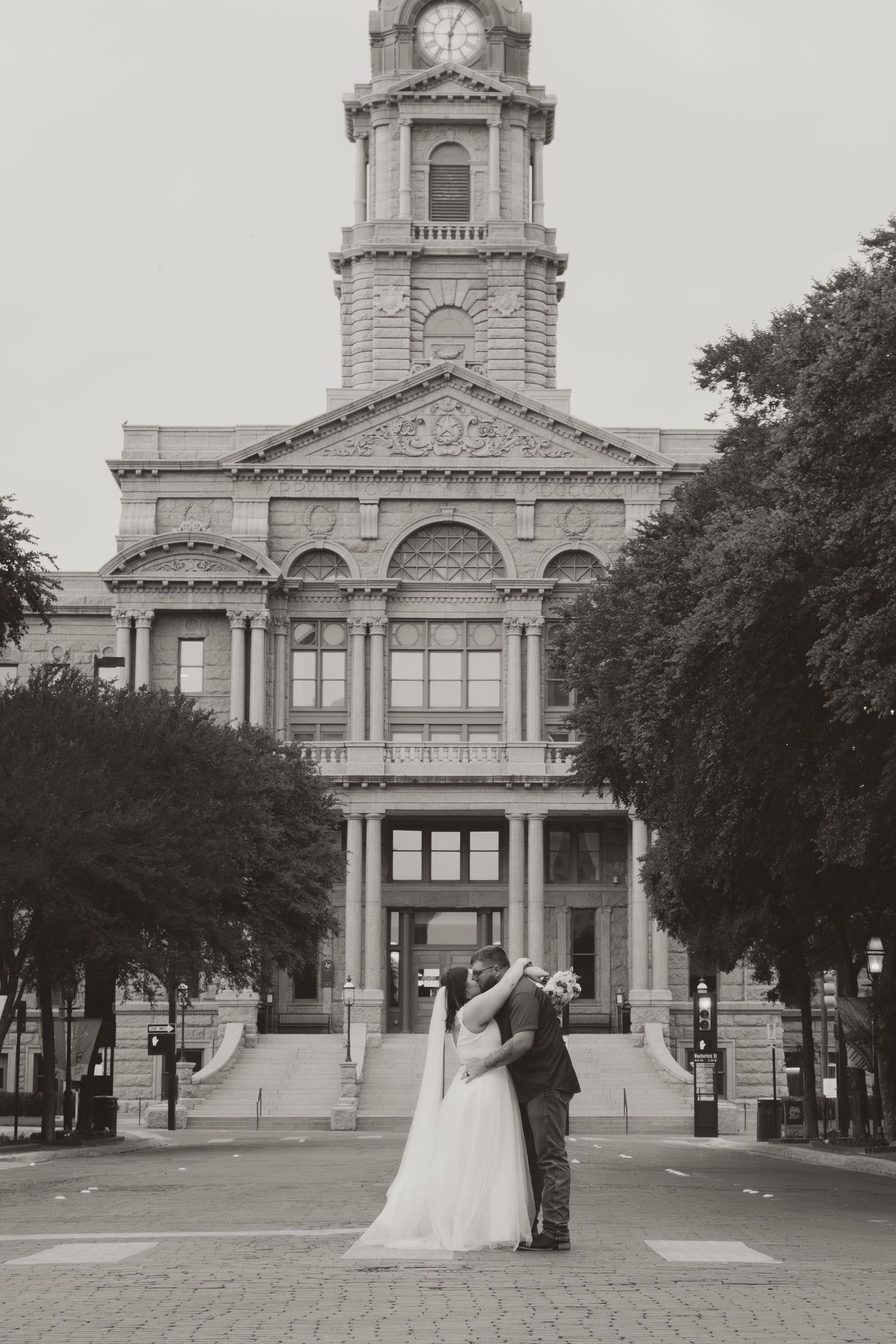 A newlywed couple shares a romantic kiss in front of a historic courthouse with a grand clock tower.