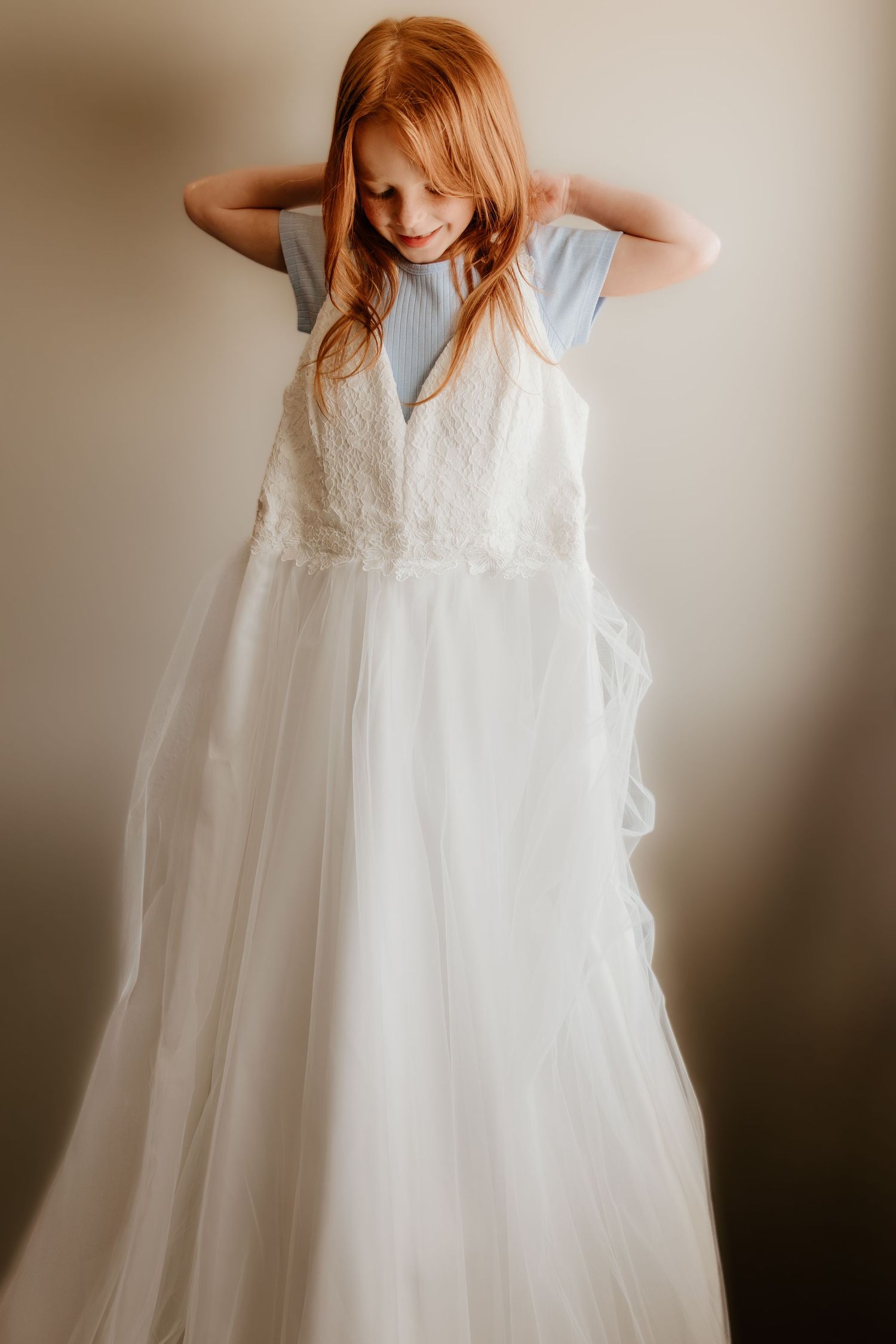 Young child wearing a flowing white formal dress poses playfully with hands behind head.