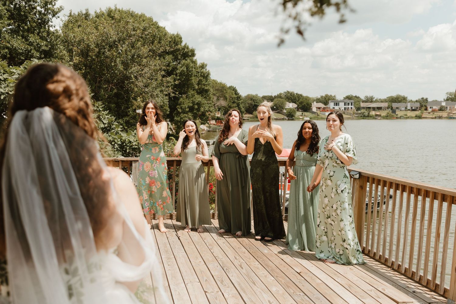 Wedding party lined up on wooden boardwalk in sage green dresses near waterfront on sunny day.