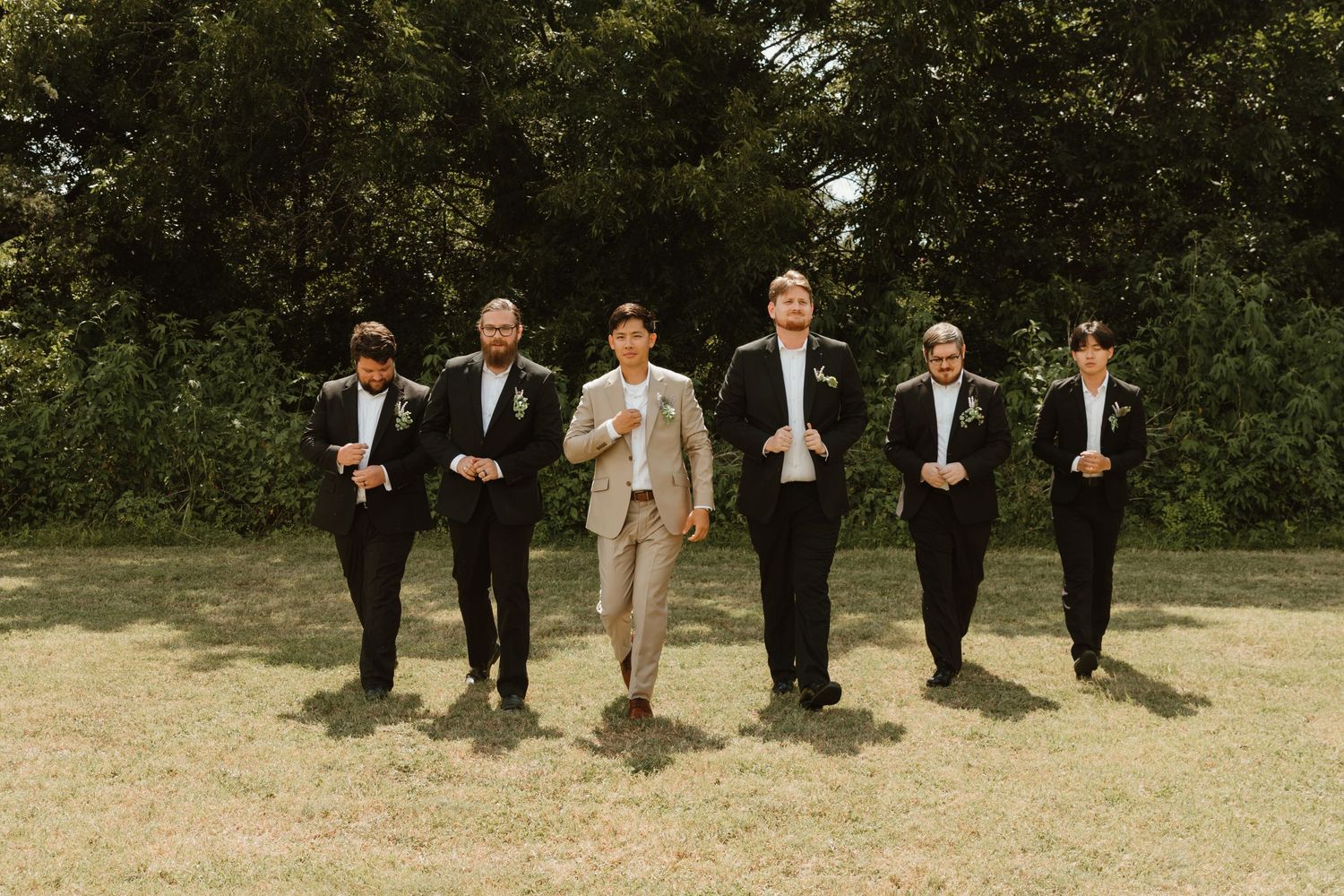 A wedding party of six people walking in a line across a grassy field with trees in the background.