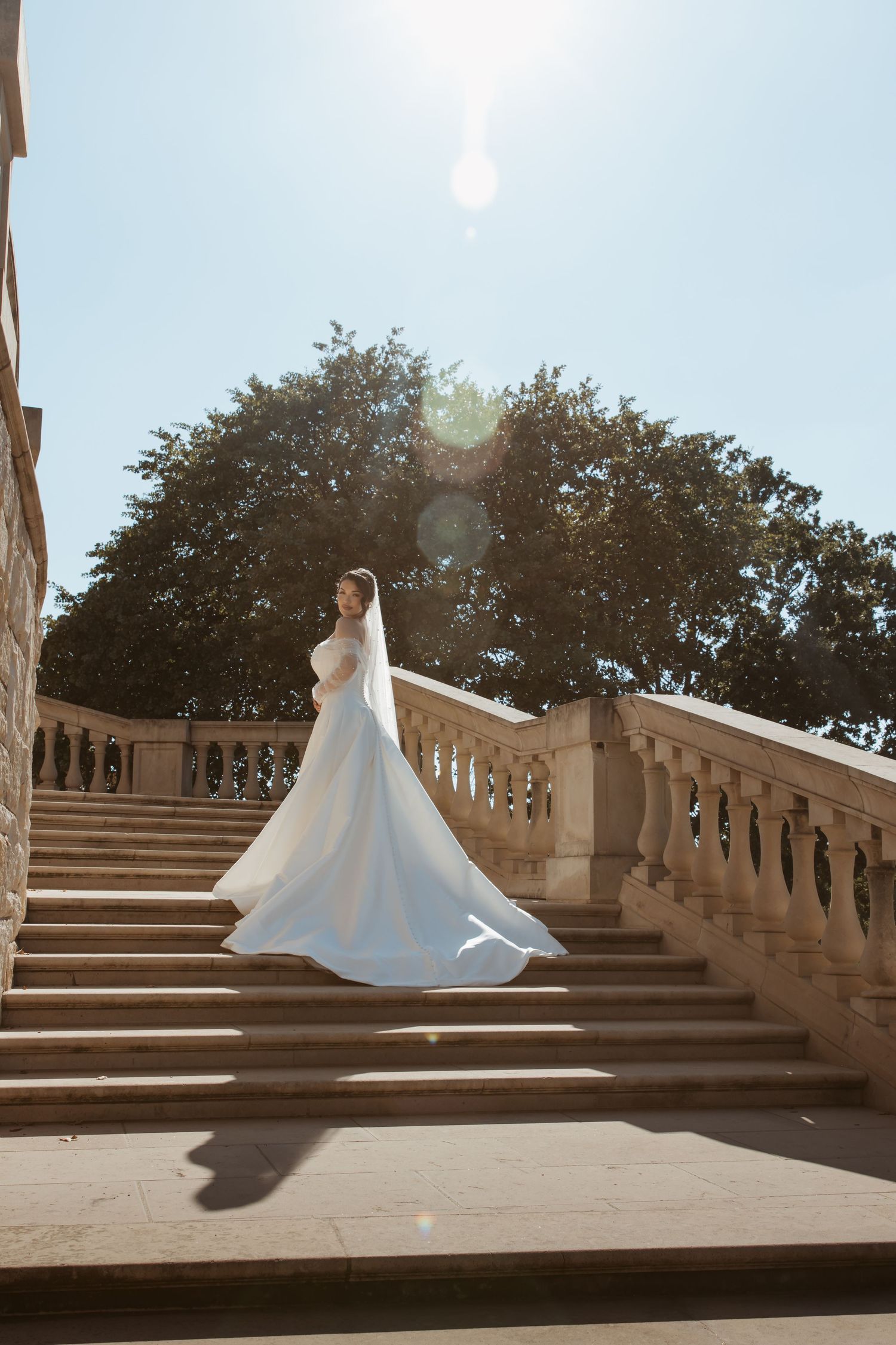A flowing white wedding dress twirls dramatically on grand stone steps against a bright sky.
