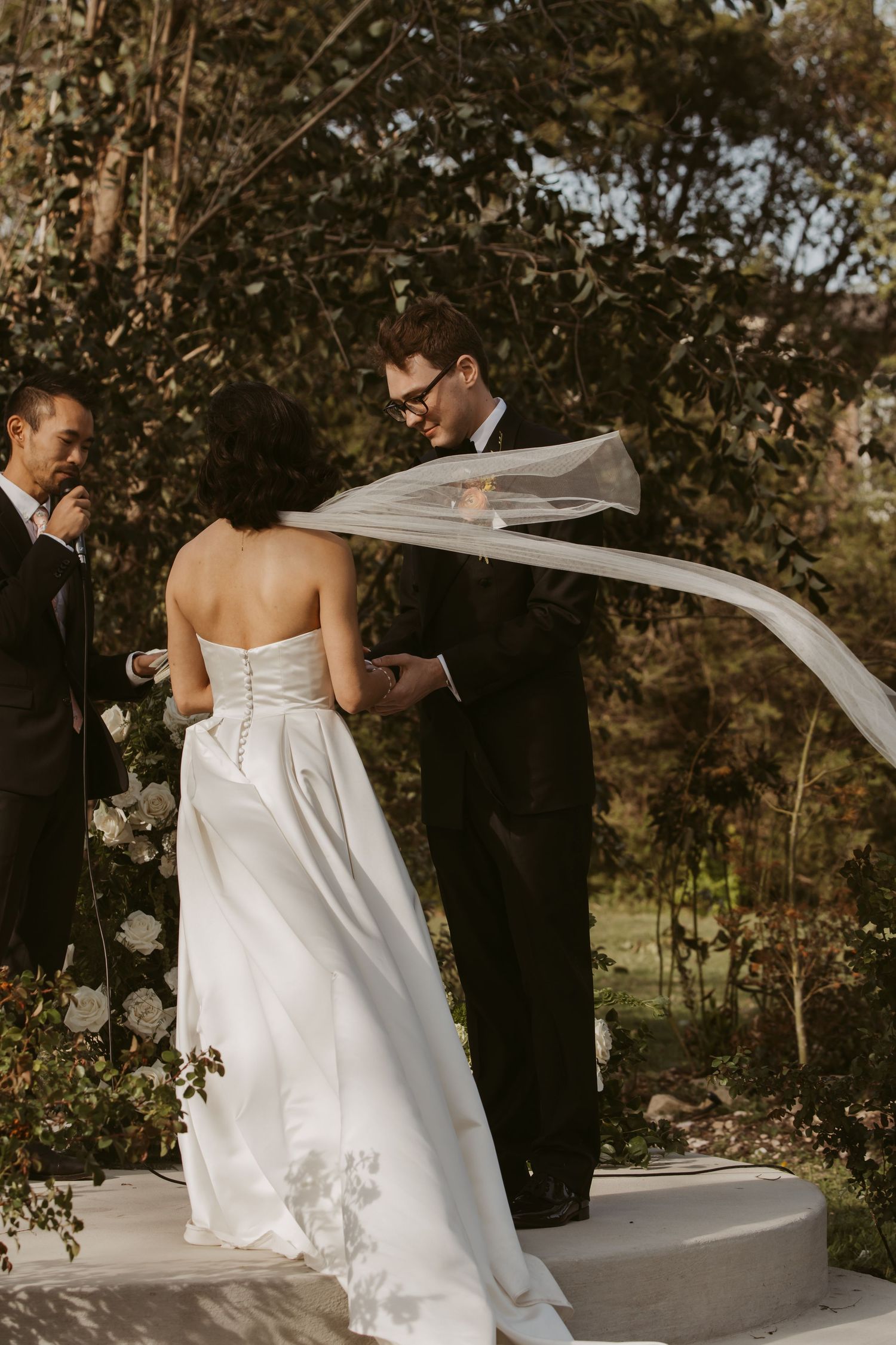 Brides veil blows in wind as groom reads vows during outdoor garden wedding ceremony.
