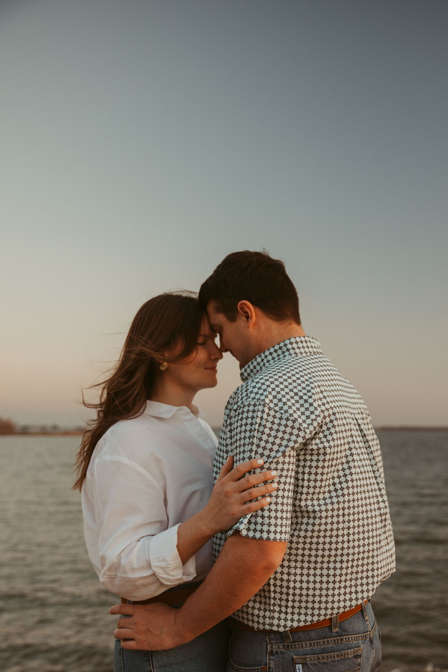 Couple embracing forehead-to-forehead at sunset by the water, woman wearing white shirt and engagement ring.