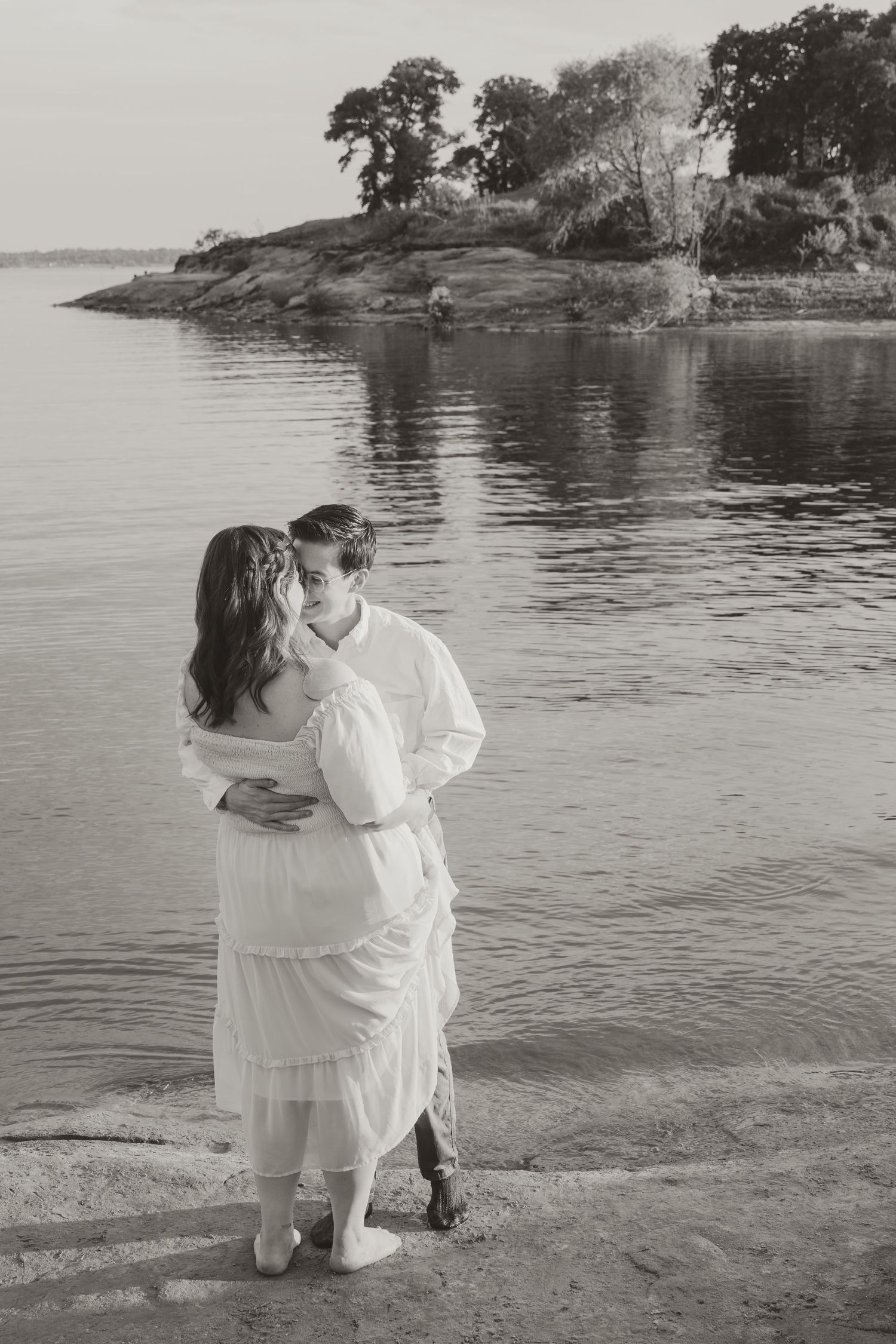 A romantic black and white photo of a couple embracing by a tranquil lake with rocky shoreline and trees.