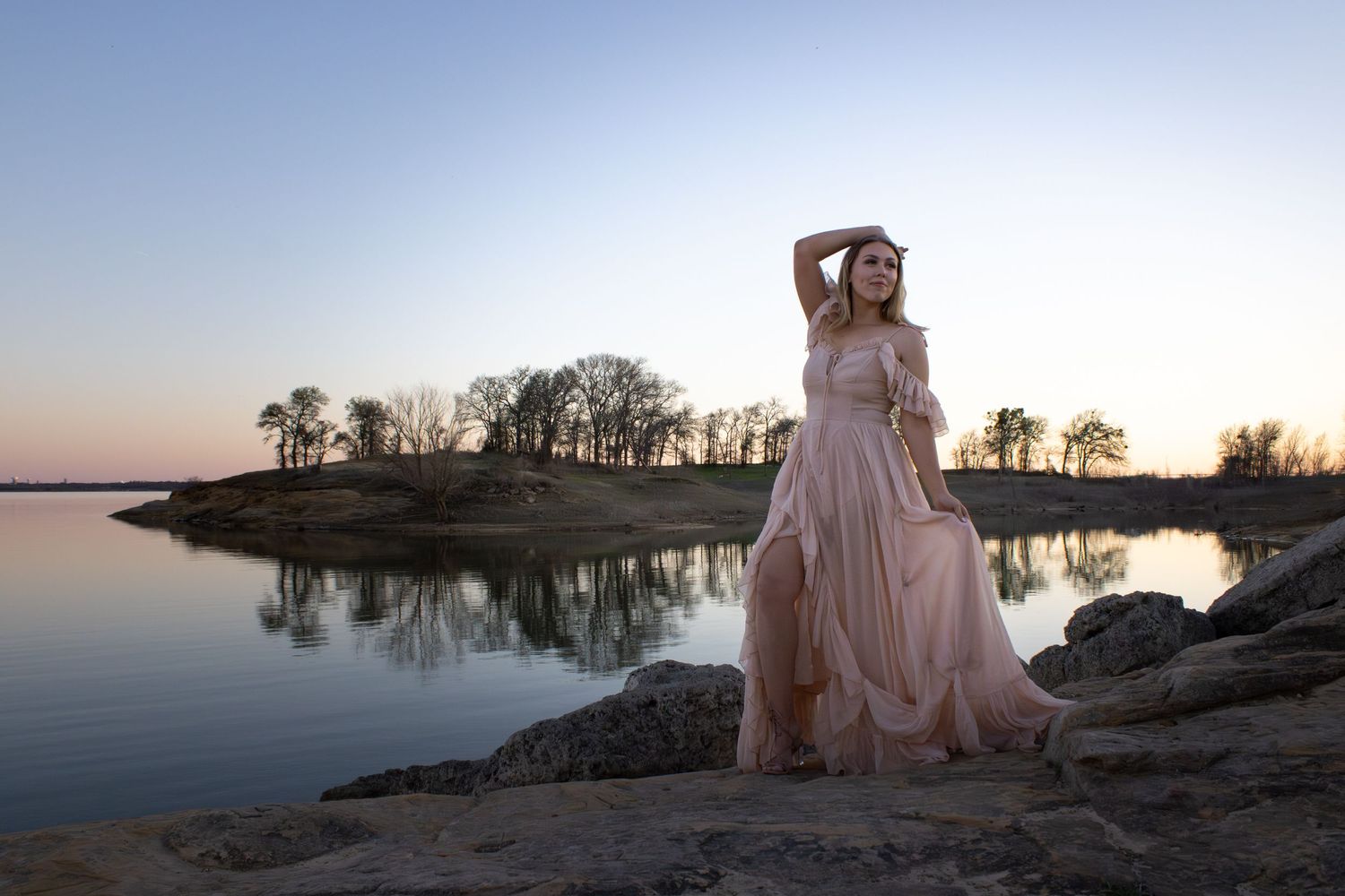 A figure in a flowing pink gown poses against a serene lakeside landscape at sunset with silhouetted trees in the background.