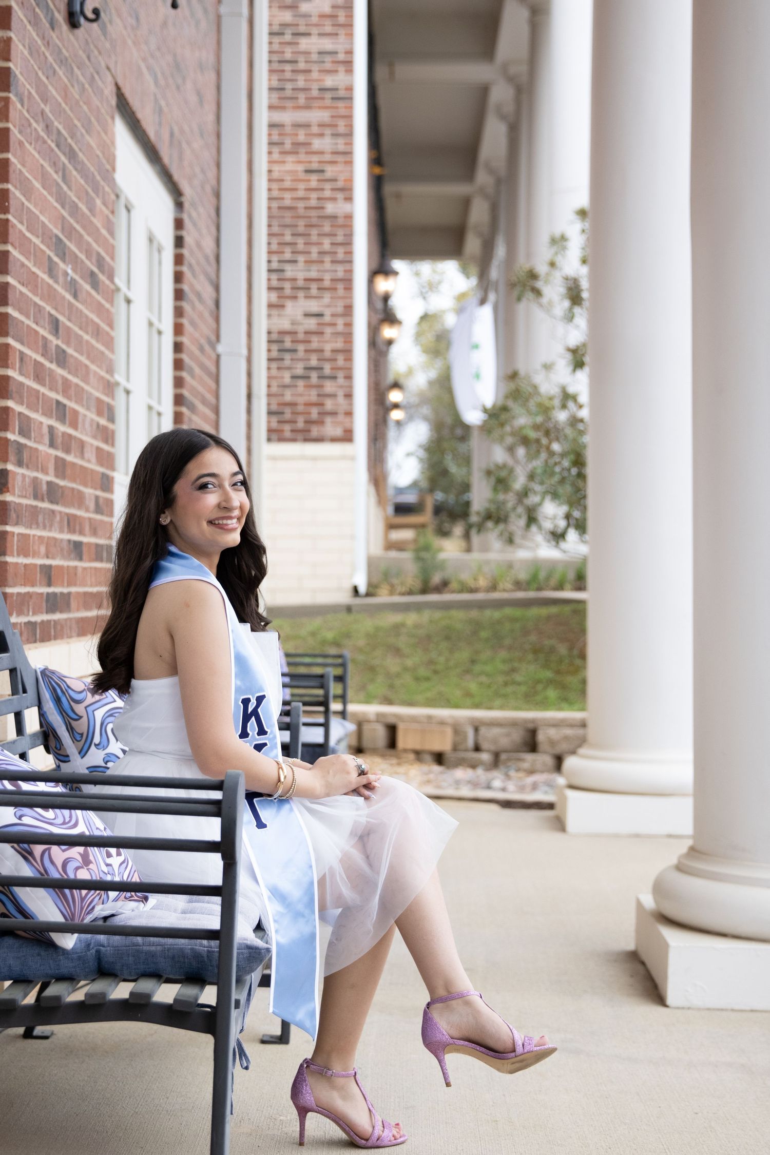 A person in a blue dress sits on a bench near brick columns and white pillars of a classical building.