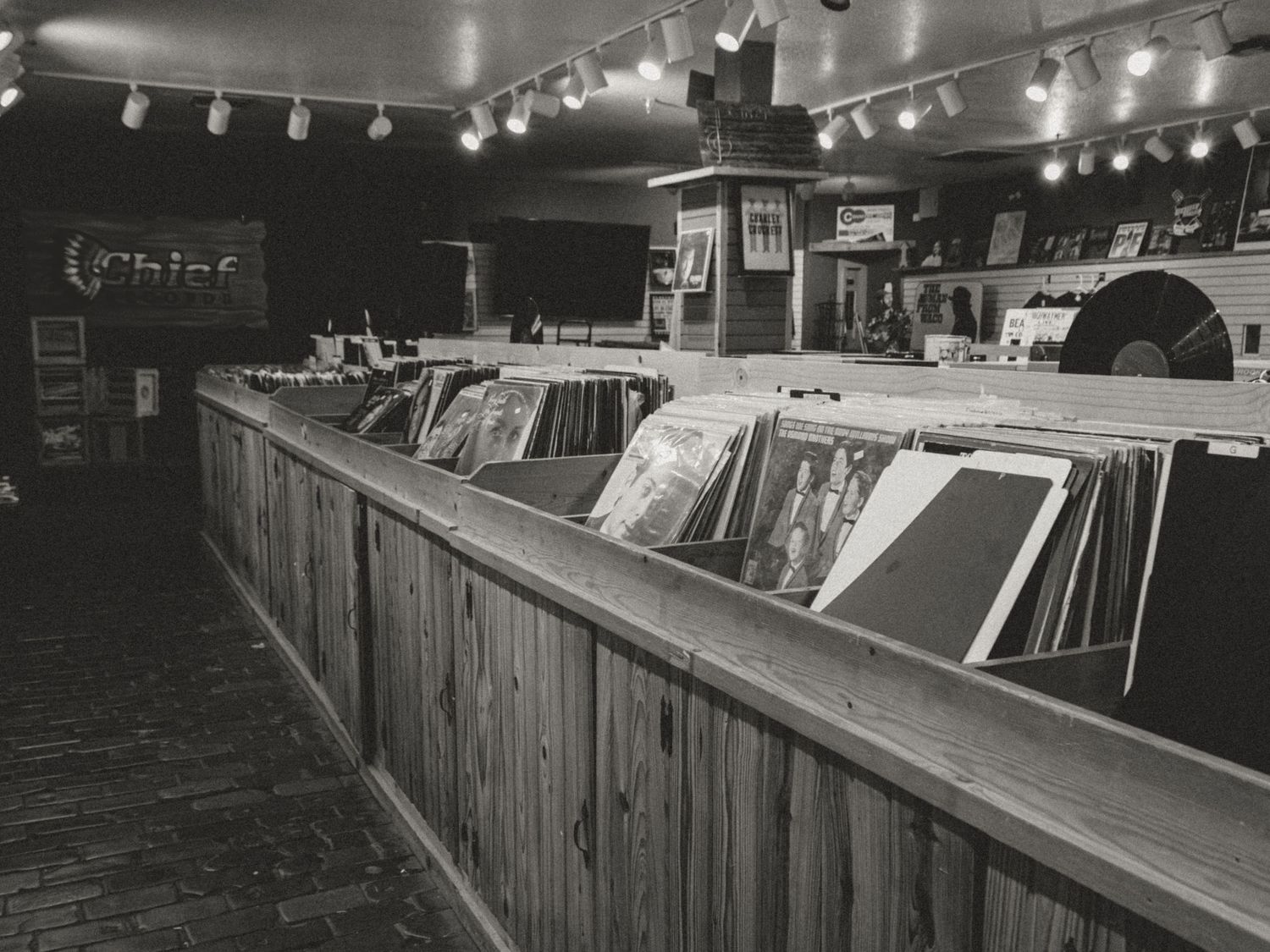 Black and white photo of vinyl records displayed in wooden bins at a vintage record store with track lighting above.