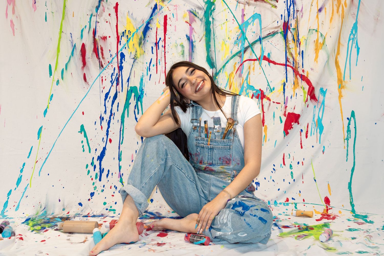Artist in paint-splattered overalls sits on white canvas floor surrounded by colorful paint splatter art.