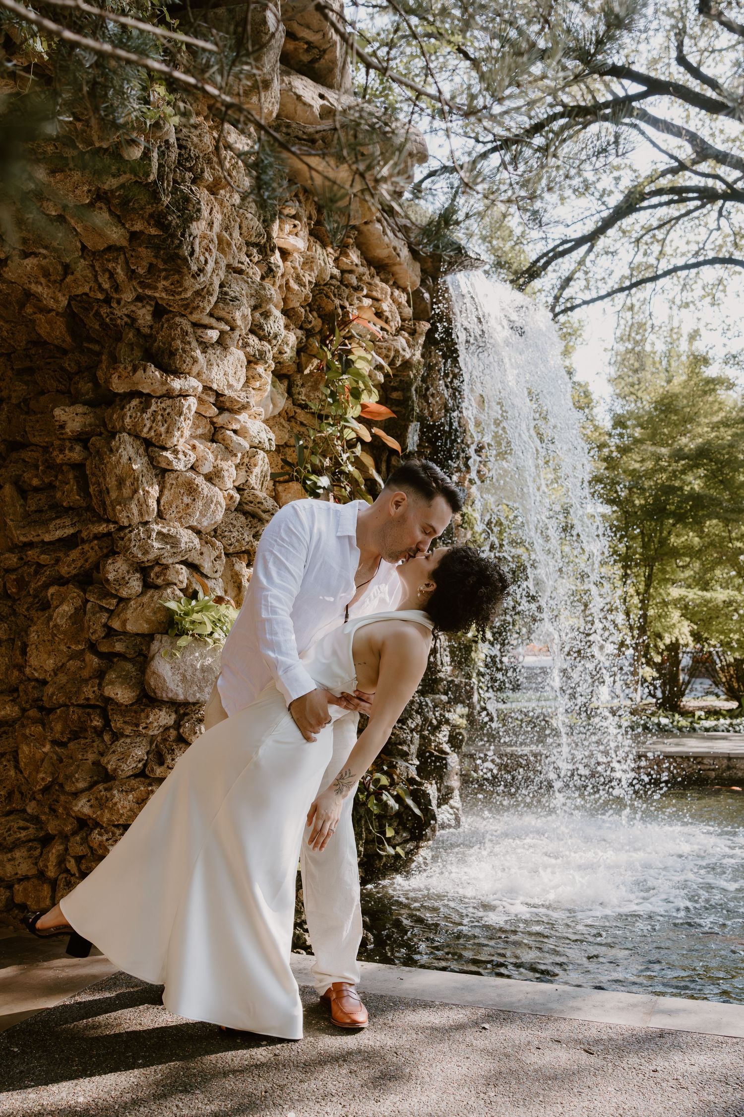 A romantic wedding photo series showing a couple in white attire posing near a cascading waterfall in a natural setting.