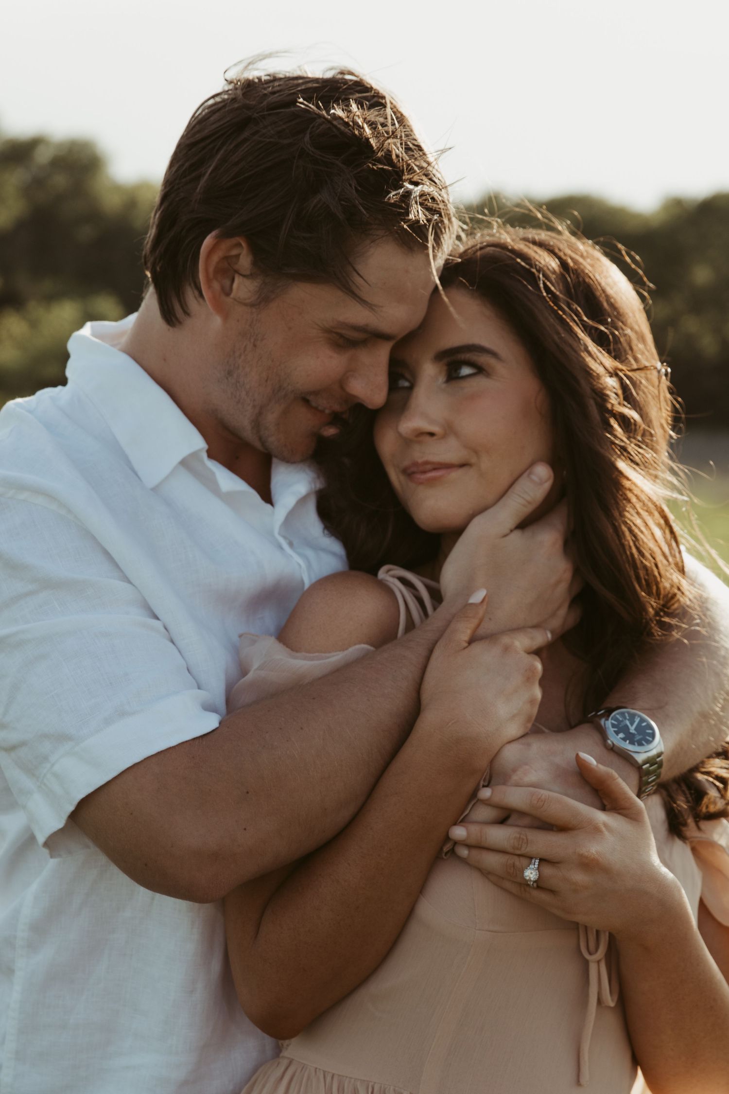 A romantic outdoor portrait of a couple embracing in soft sunlight, with her wearing a blush dress and him in a white shirt.