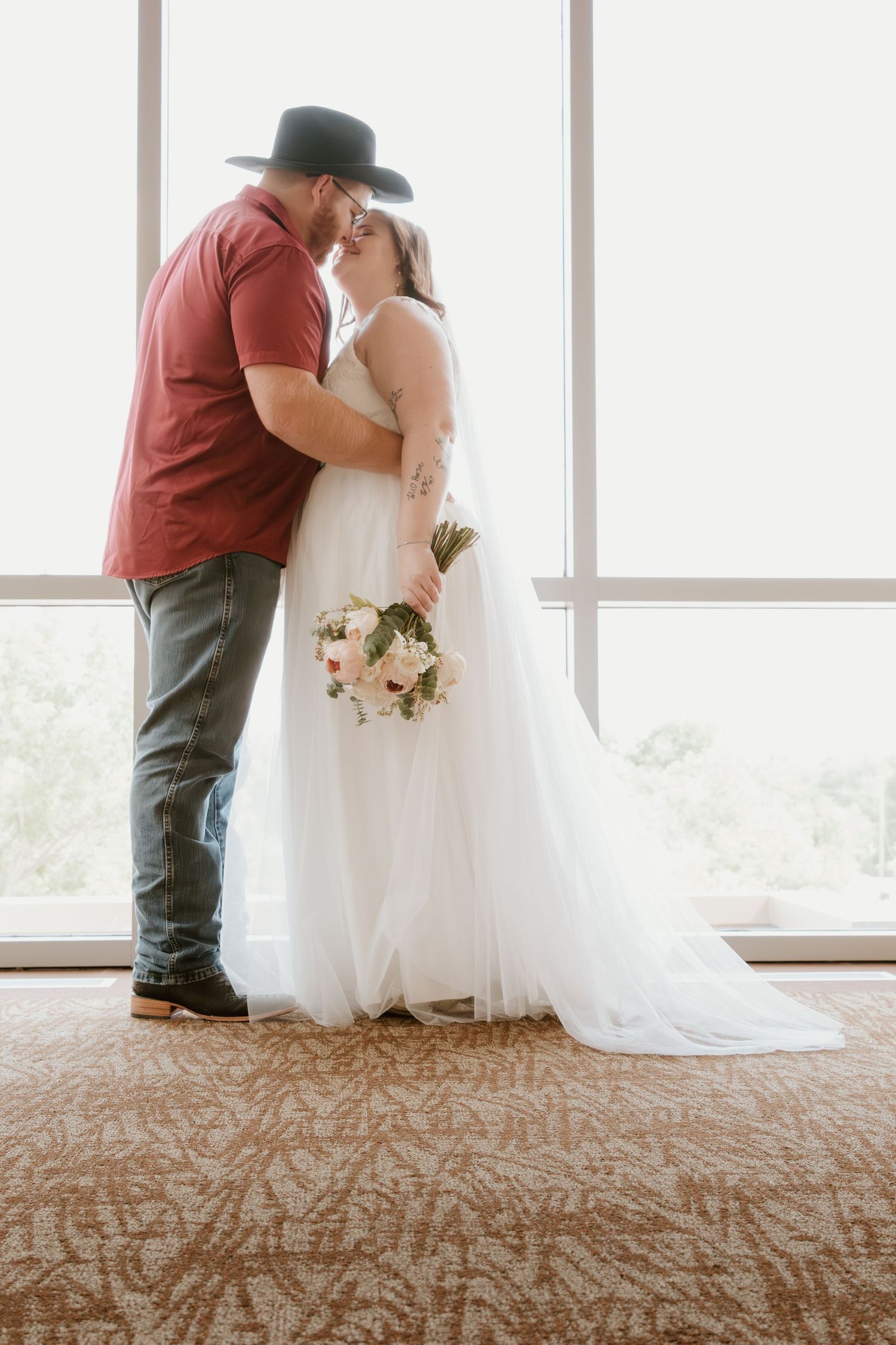 Couple in wedding attire sharing romantic moment near large windows.