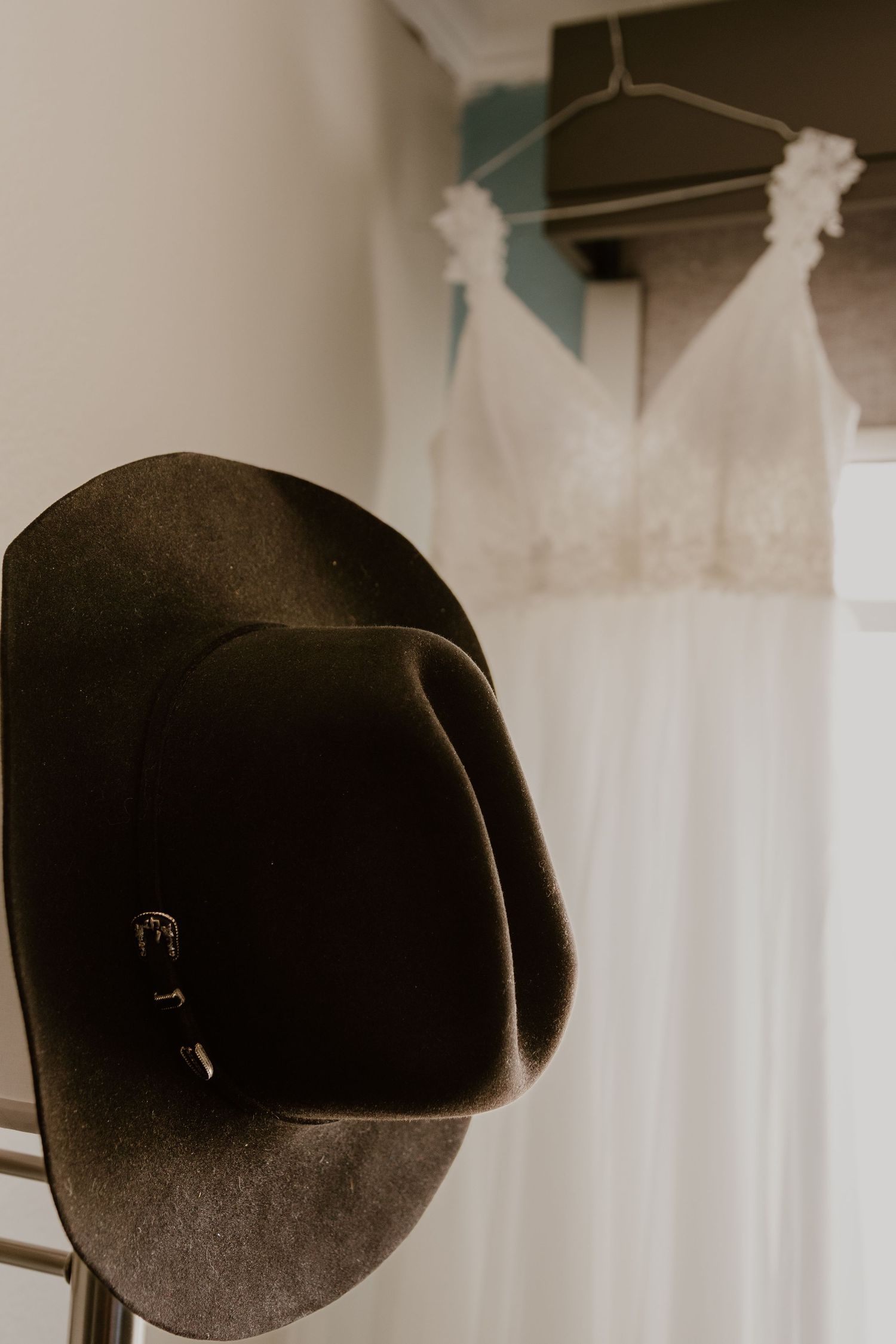 A cowboy hat and white wedding dress hanging side by side against a light colored wall.