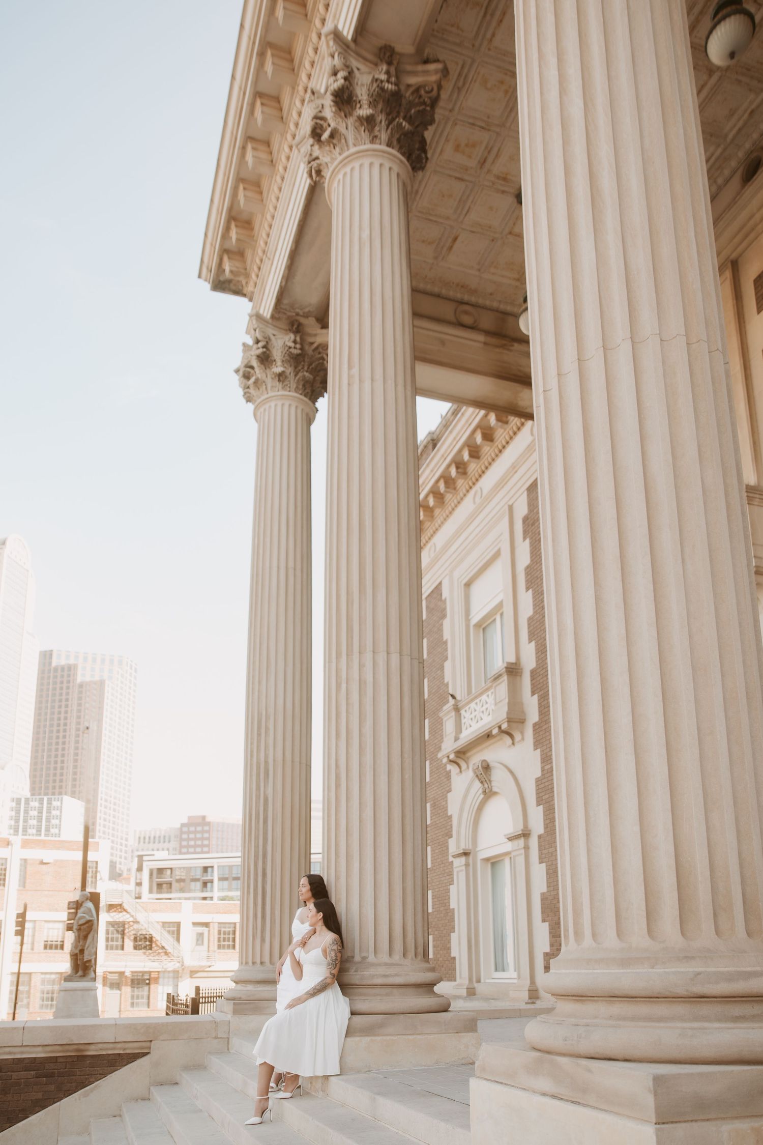 Woman in white dress standing beside tall classical columns of a grand historic building in an urban setting.