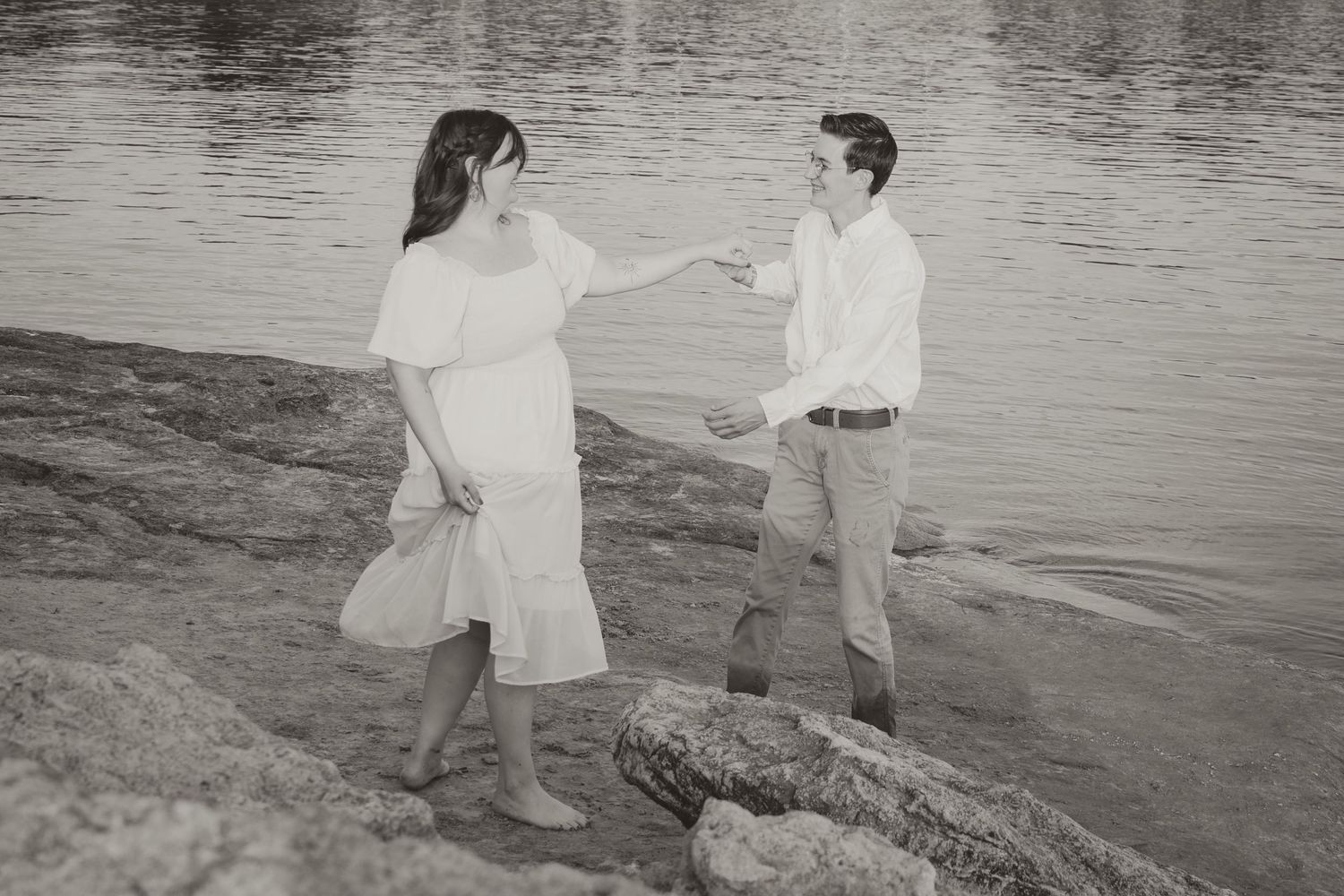 A couple in white outfits shares a romantic moment by the water's edge in a black and white photograph.