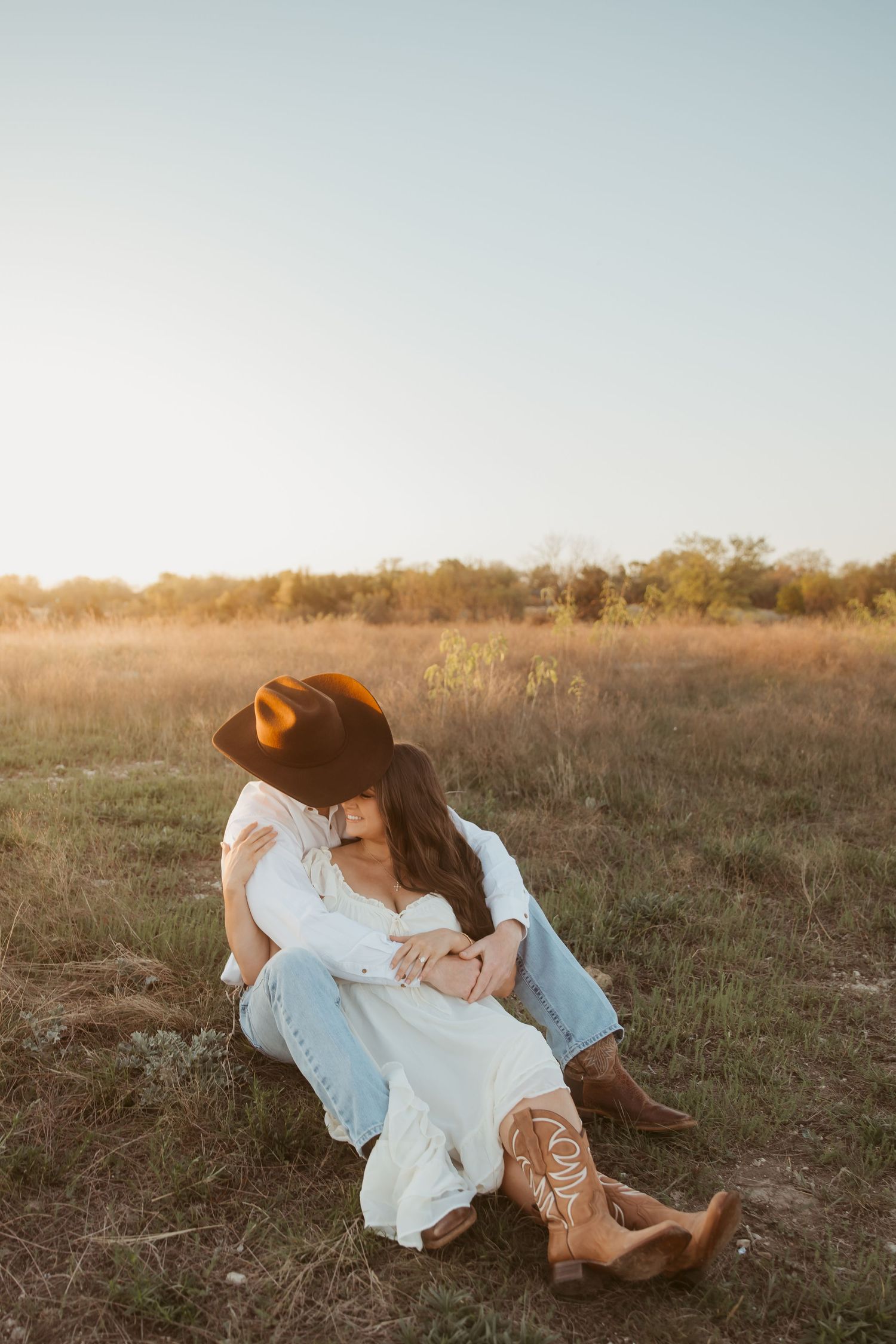 Couple sitting together in a sunlit field, woman in white dress and cowboy boots, man in cowboy hat.