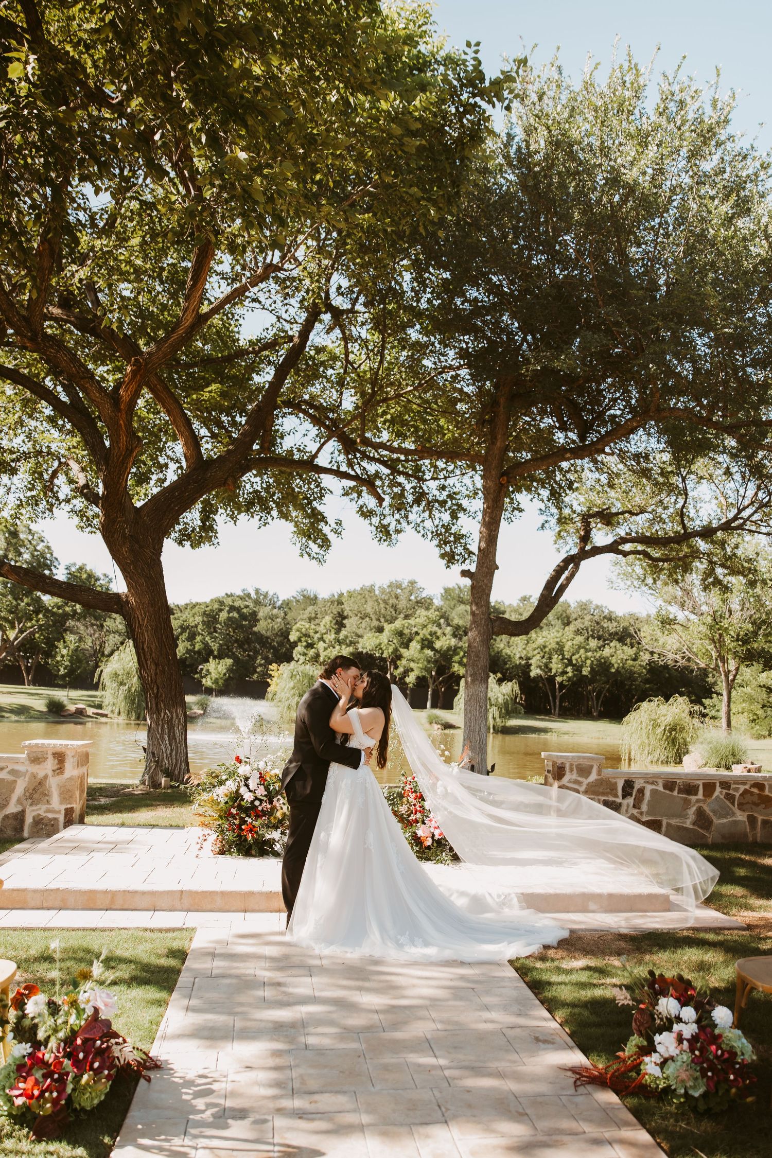 Bride and groom share first kiss at outdoor wedding ceremony under large oak trees with floral arrangements and flowing veil.