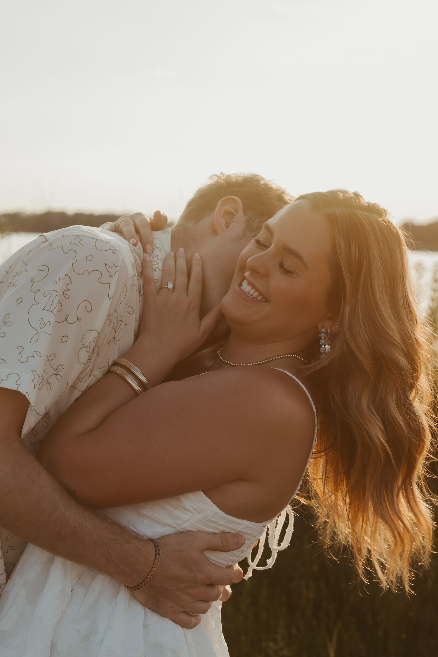 Couple embracing at golden hour sunset, woman in white dress smiling joyfully, warm romantic outdoor engagement photo.