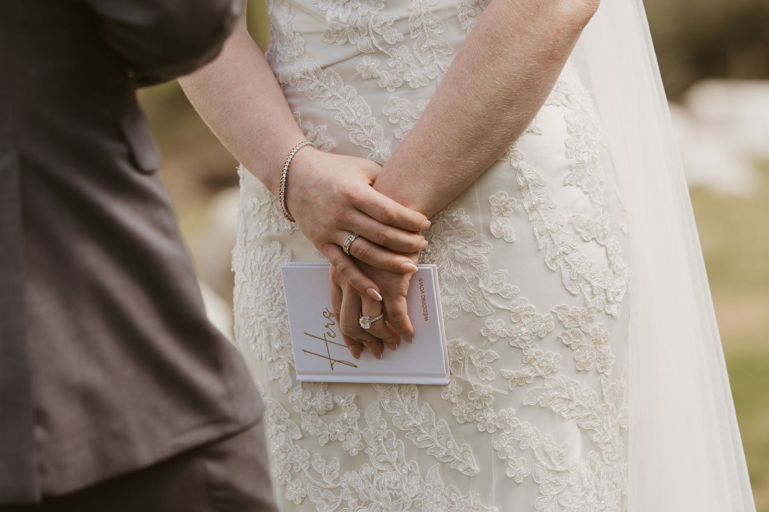 Bride and groom holding hands during wedding ceremony, bride wearing lace gown and diamond ring, holding vows booklet.