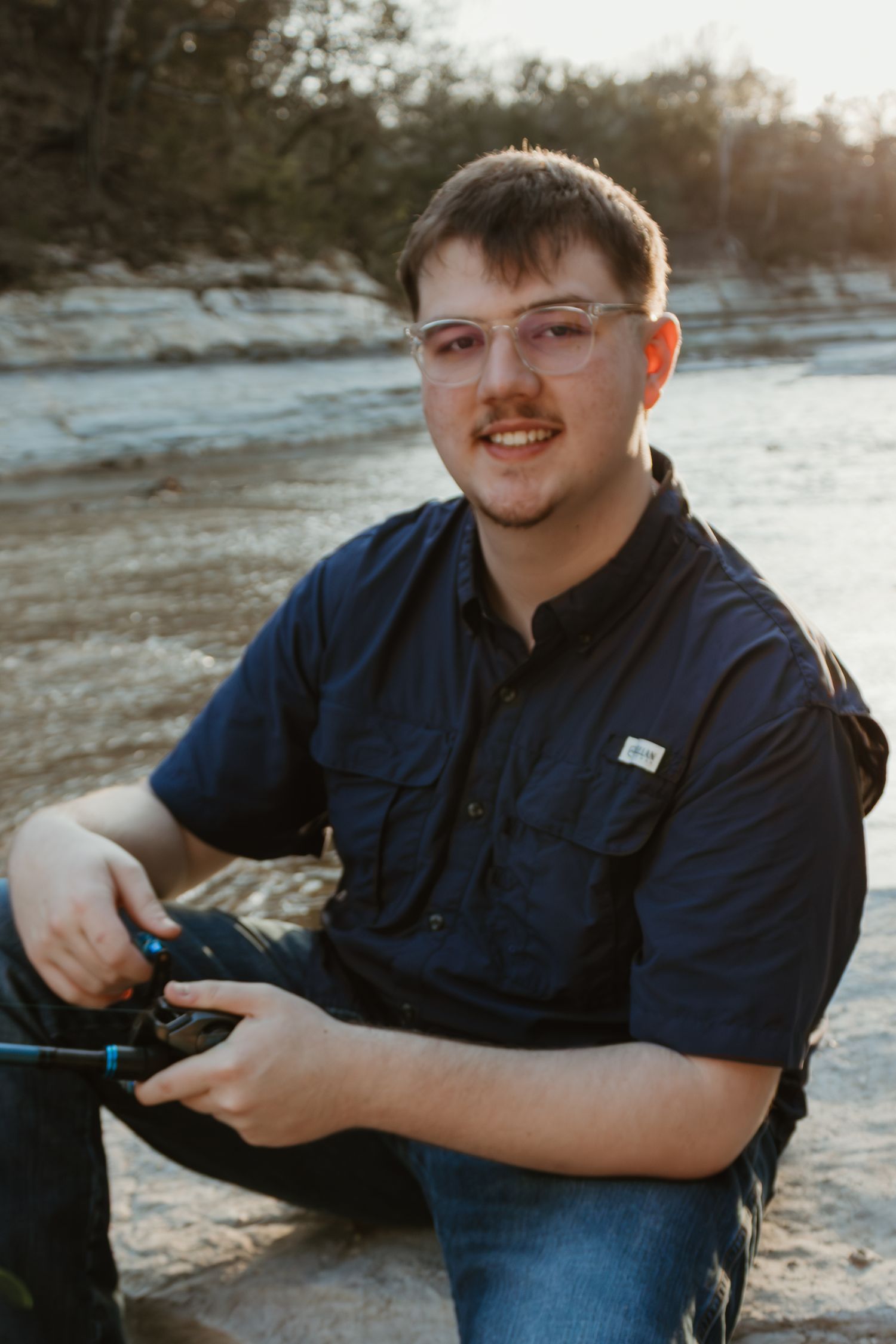 A person in a navy blue shirt and glasses sits outdoors near a rocky shoreline at sunset.