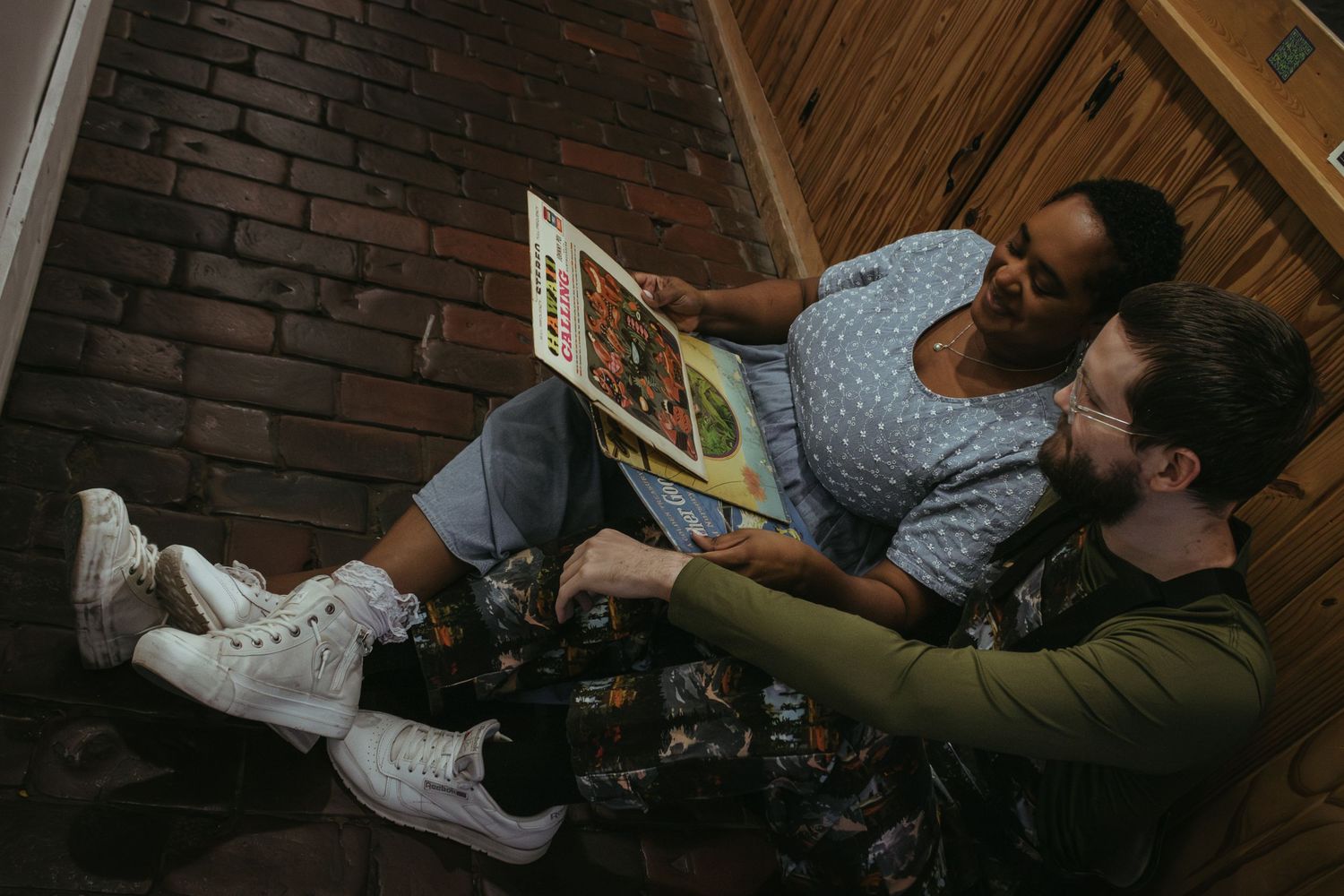 Two people sit close together on wooden floor against brick wall while reading a book together.