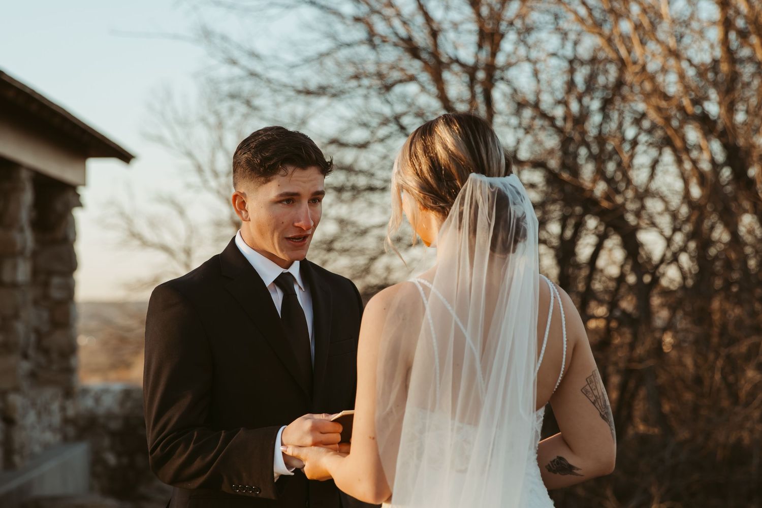 A couple shares an intimate moment during their wedding ceremony at sunset with bare trees in the background.