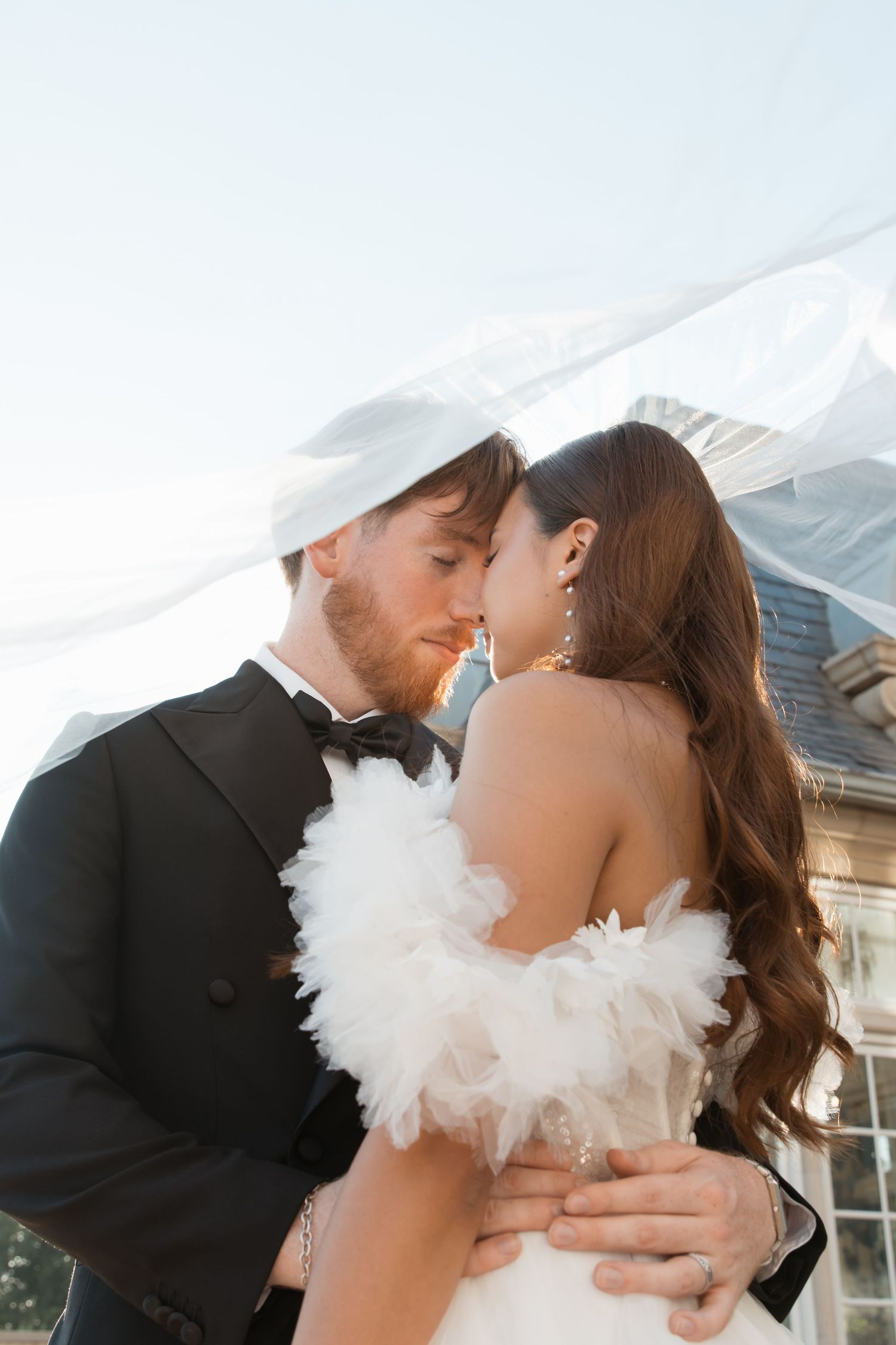 A romantic wedding couple embraces while wearing formal attire against a mountain backdrop.