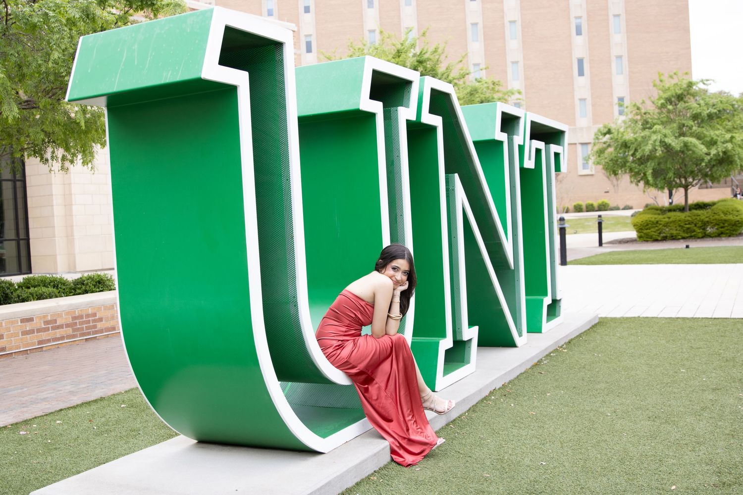 A large green UNT (University of North Texas) letter sculpture sits on grass with someone in a red dress resting against it.