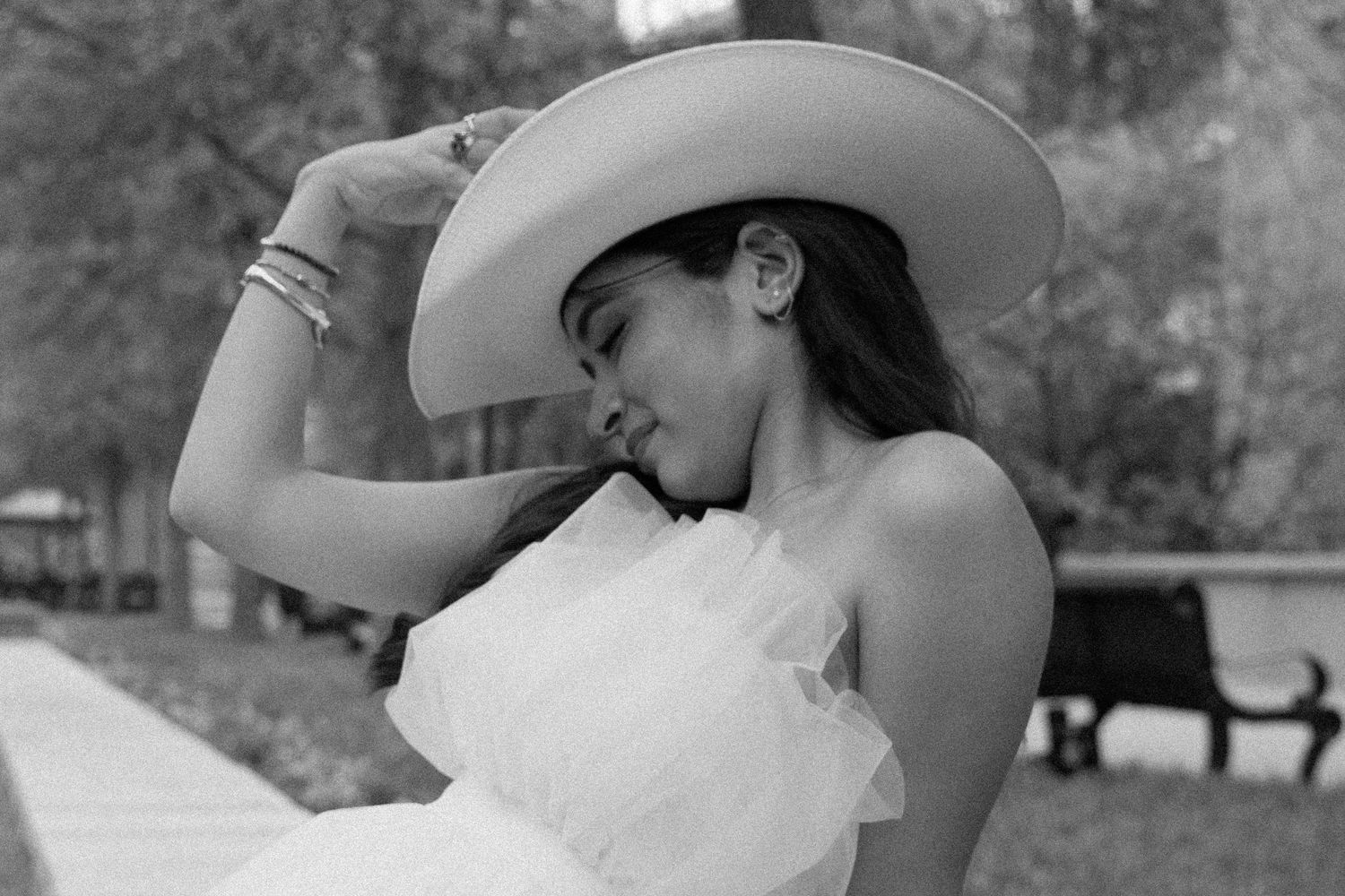 A person in a white strapless dress and cowboy hat poses outdoors in black and white photography.