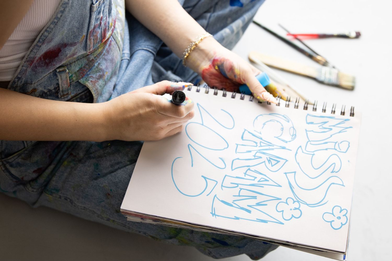 Someone sketches blue bubble letters in a spiral notebook with markers while sitting at a desk.