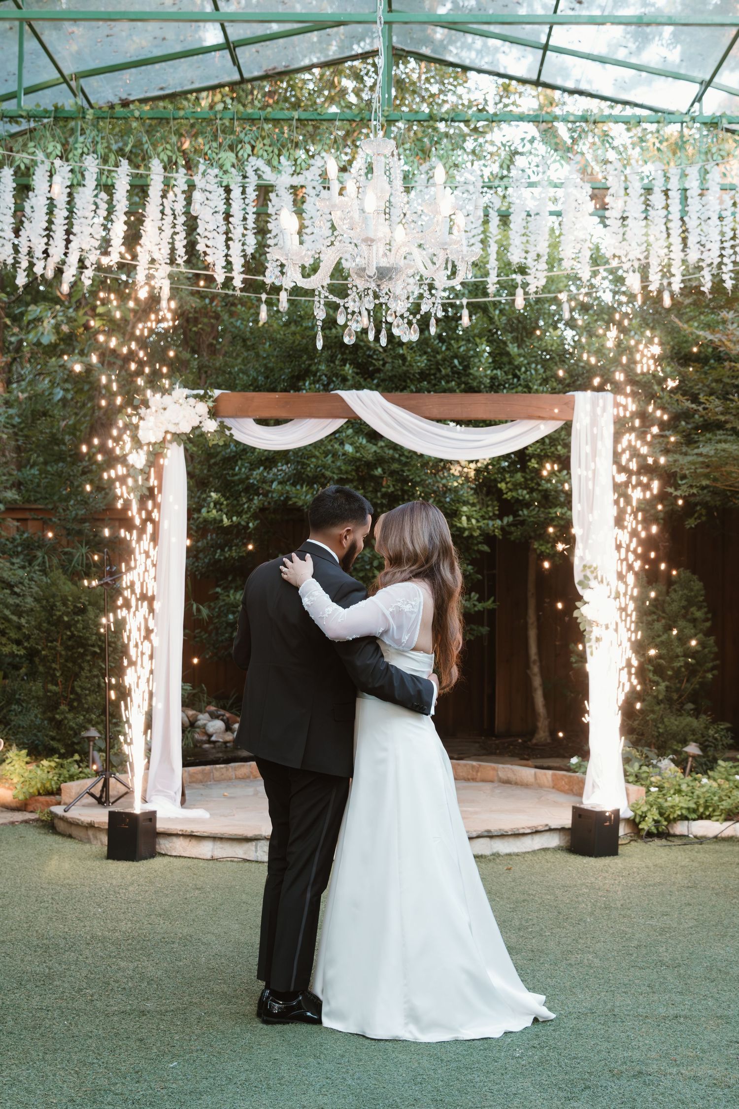 Wedding couple twirling under hanging floral decorations and string lights in a garden reception setting.