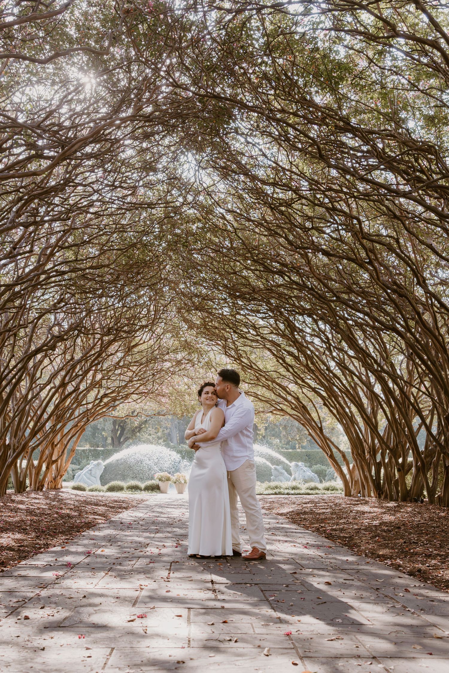 A couple embraces under a natural archway of curved tree branches creating a romantic tunnel effect on a sunny day.