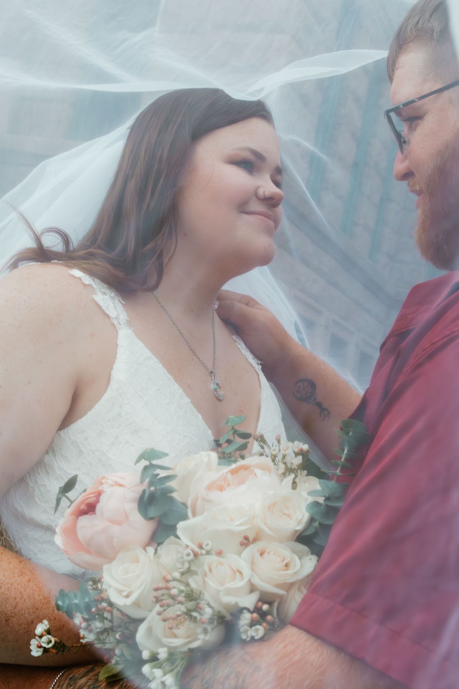 A tender moment between newlyweds captured through a flowing veil with bouquet of pink and white roses.