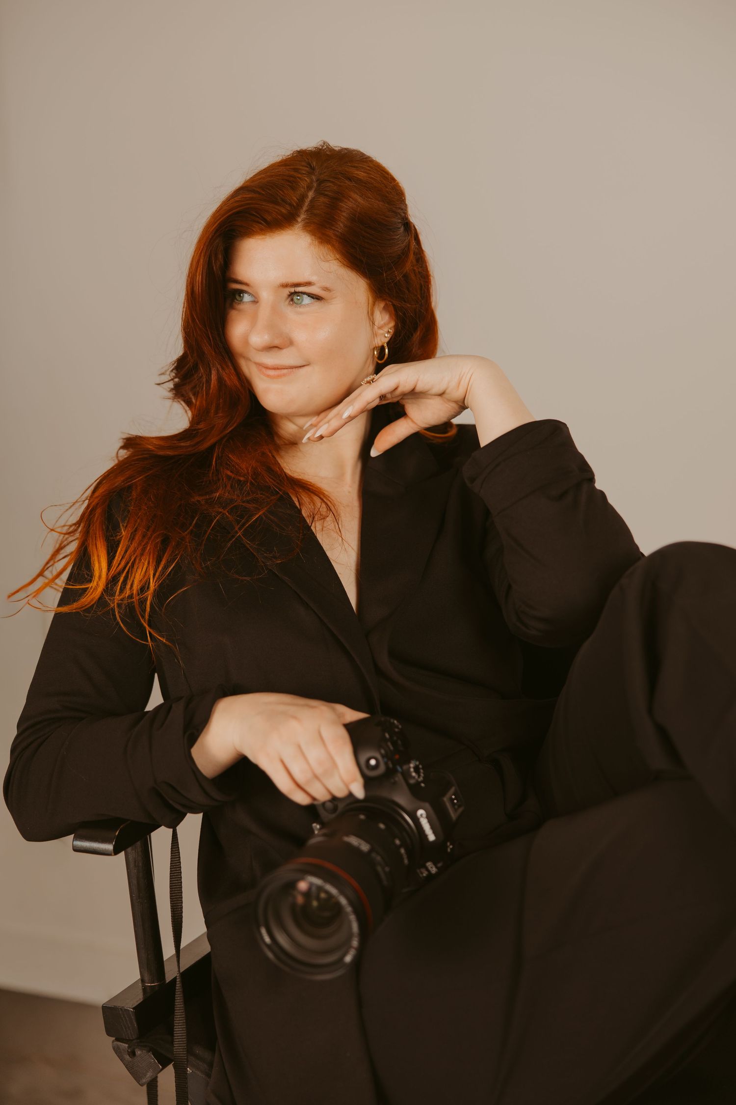A photographer in a black blazer poses with a professional camera against a neutral studio backdrop.