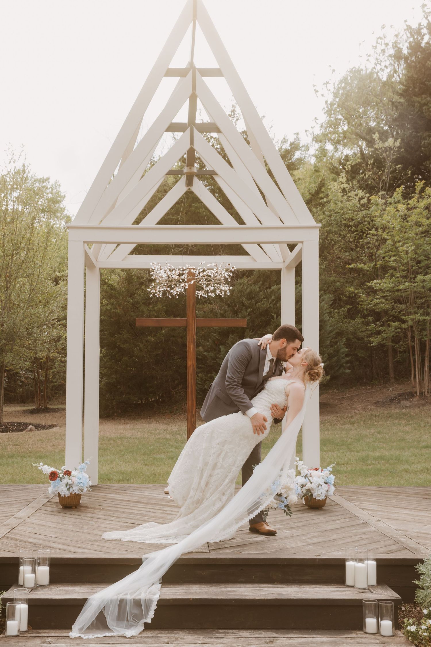 Bride and groom share a romantic dip kiss under a white wooden chapel arch with a cross and floral chandelier outdoors.