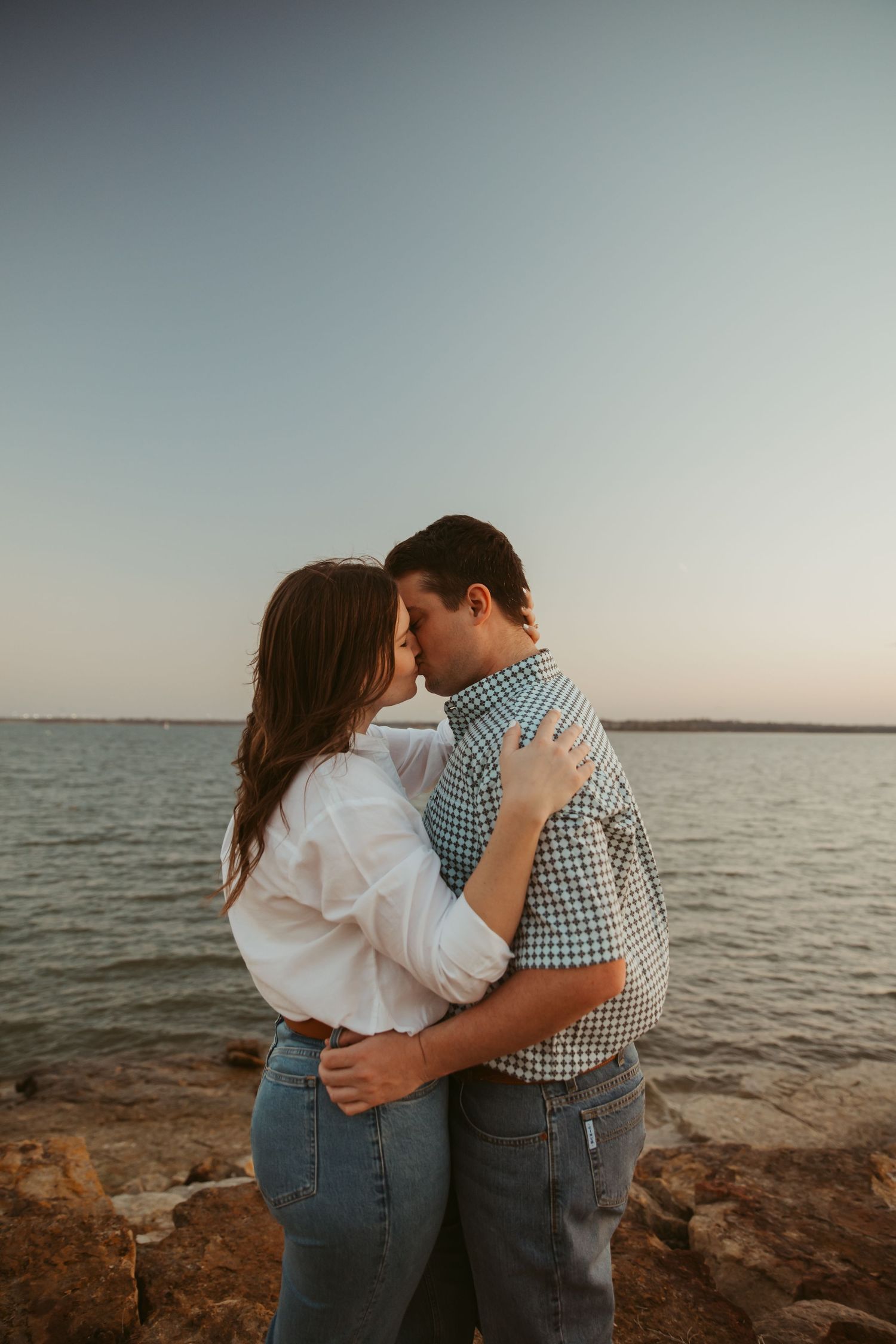 Couple sharing a romantic kiss by the water at golden hour, embracing on rocky shoreline at sunset.