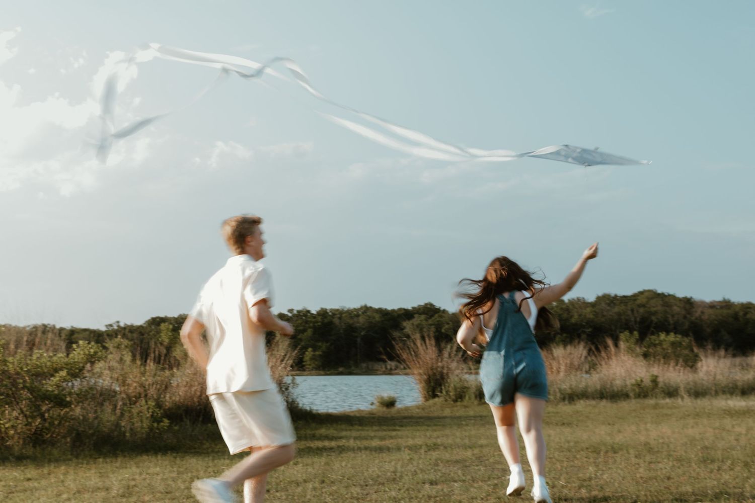 Two people dance and celebrate on a grassy field near a lake with mountains in the background on a cloudy day.